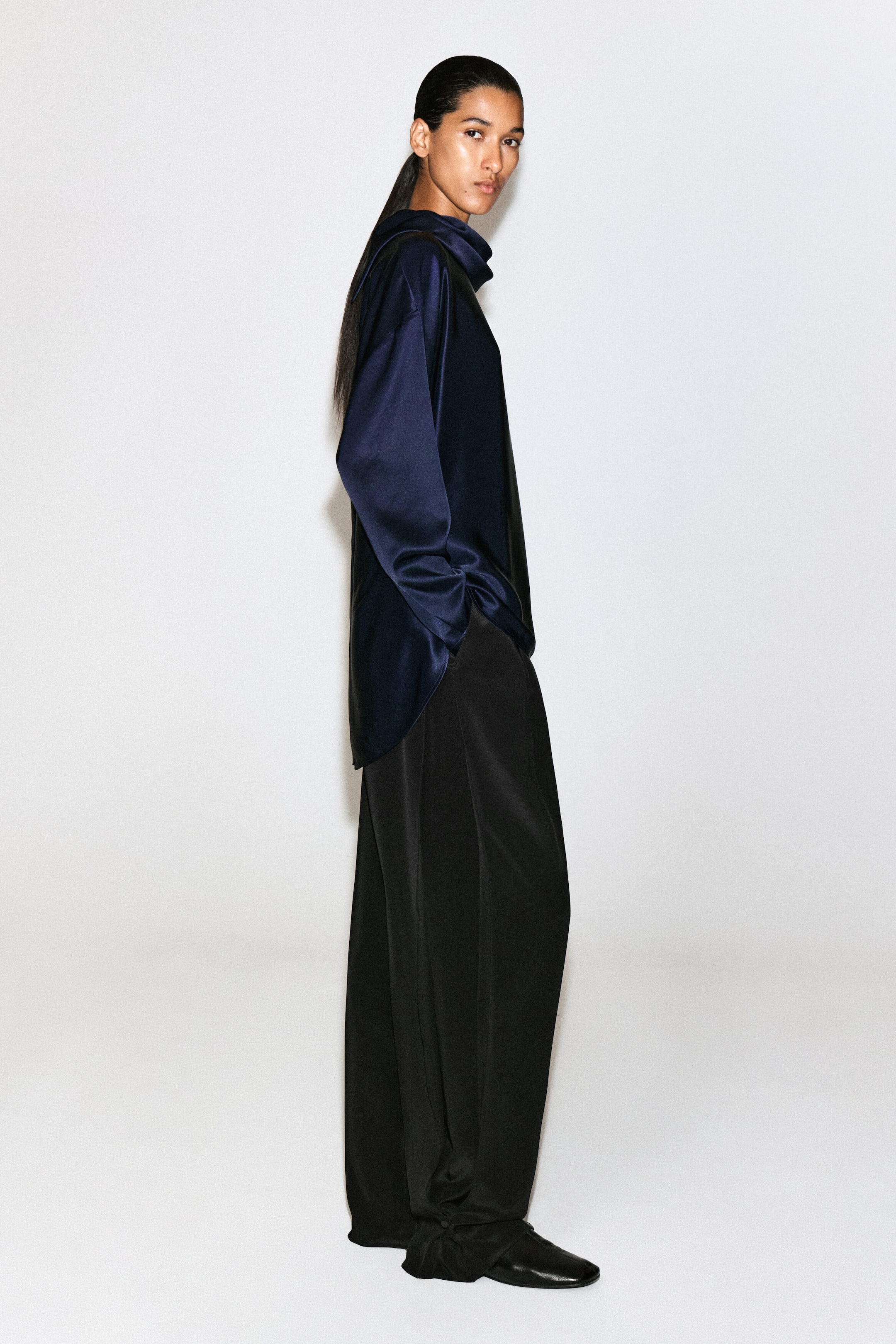 A navy blue cowl-neck satin blouse is shown in side profile facing right, highlighting its relaxed fit and lustrous draped fabric. The blouse features long sleeves and a high, folded cowl neck, with its curved hemline falling loosely over black wide-leg trousers.