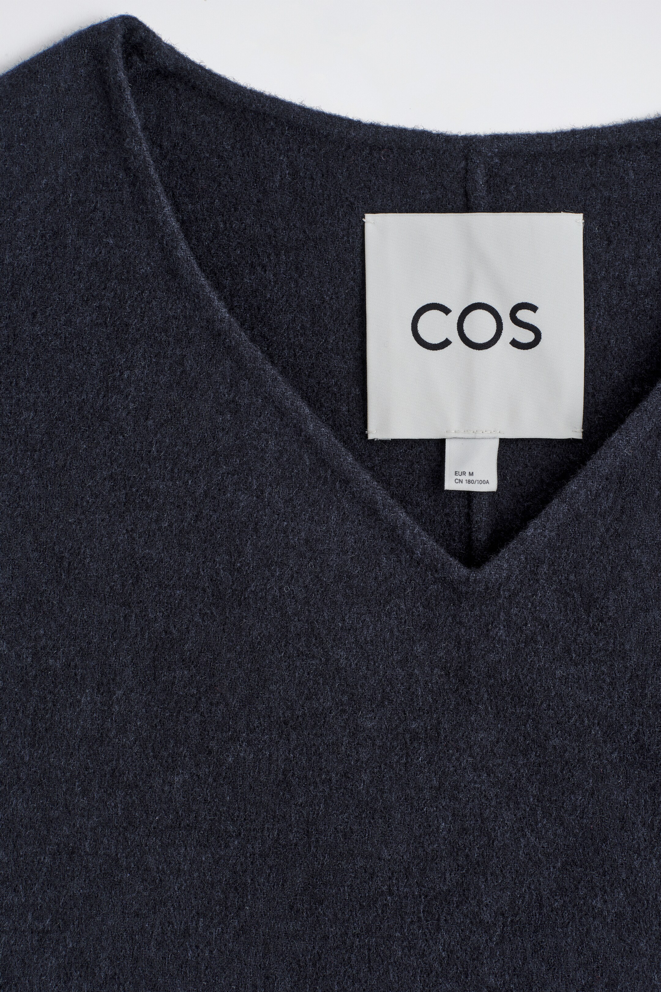 Agrandir l'image: Un gilet bleu marine à col en V présente une texture douce et brossée ainsi qu'un profond décolleté en V, avec une étiquette de marque blanche 'COS' visible en son centre.