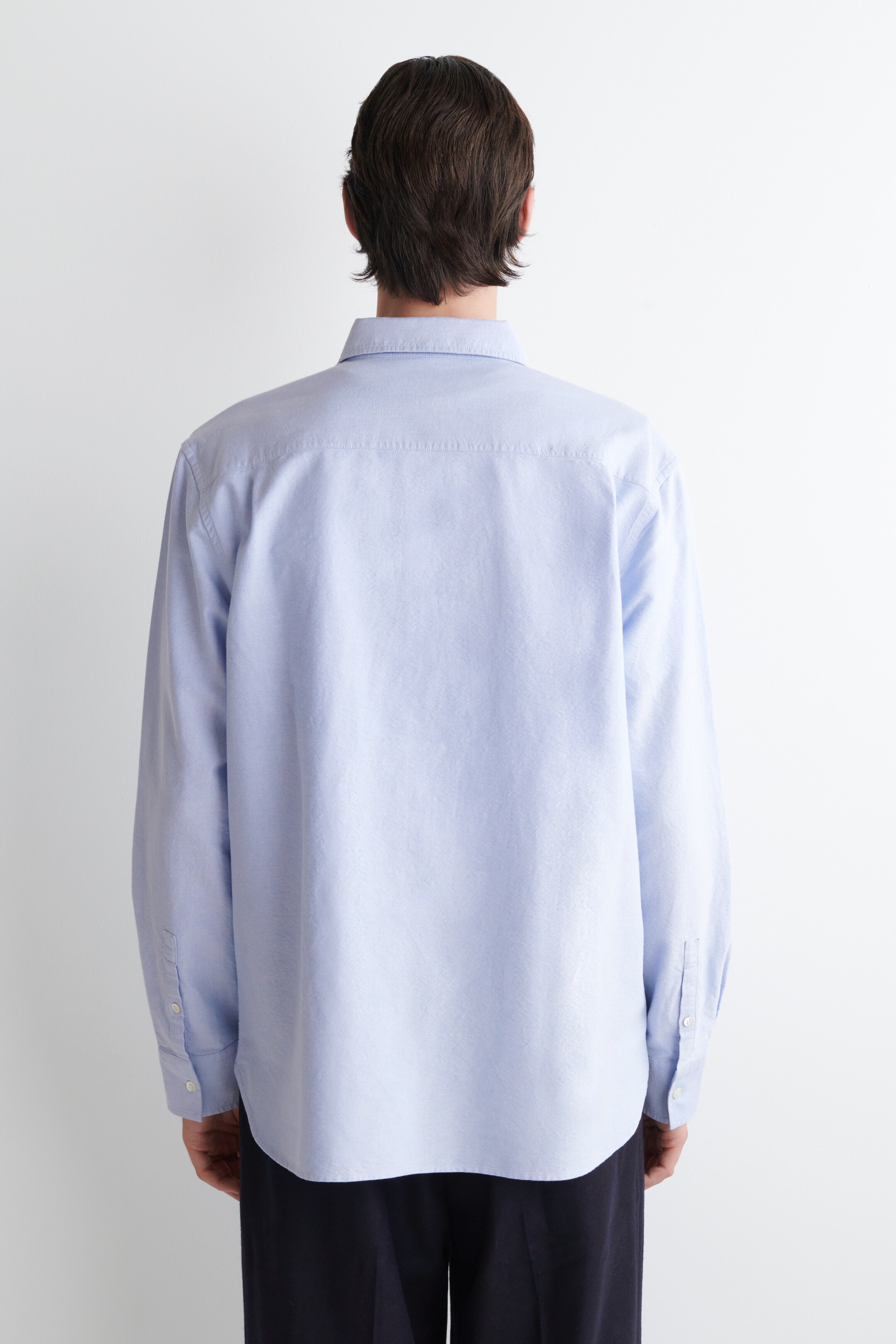 Ver imagen más grande: Una camisa de manga larga azul claro se está viendo desde atrás, con un cuello clásico, un canesú trasero visible y puños abotonados. La camisa presenta un ajuste relajado con un dobladillo suavemente curvado.