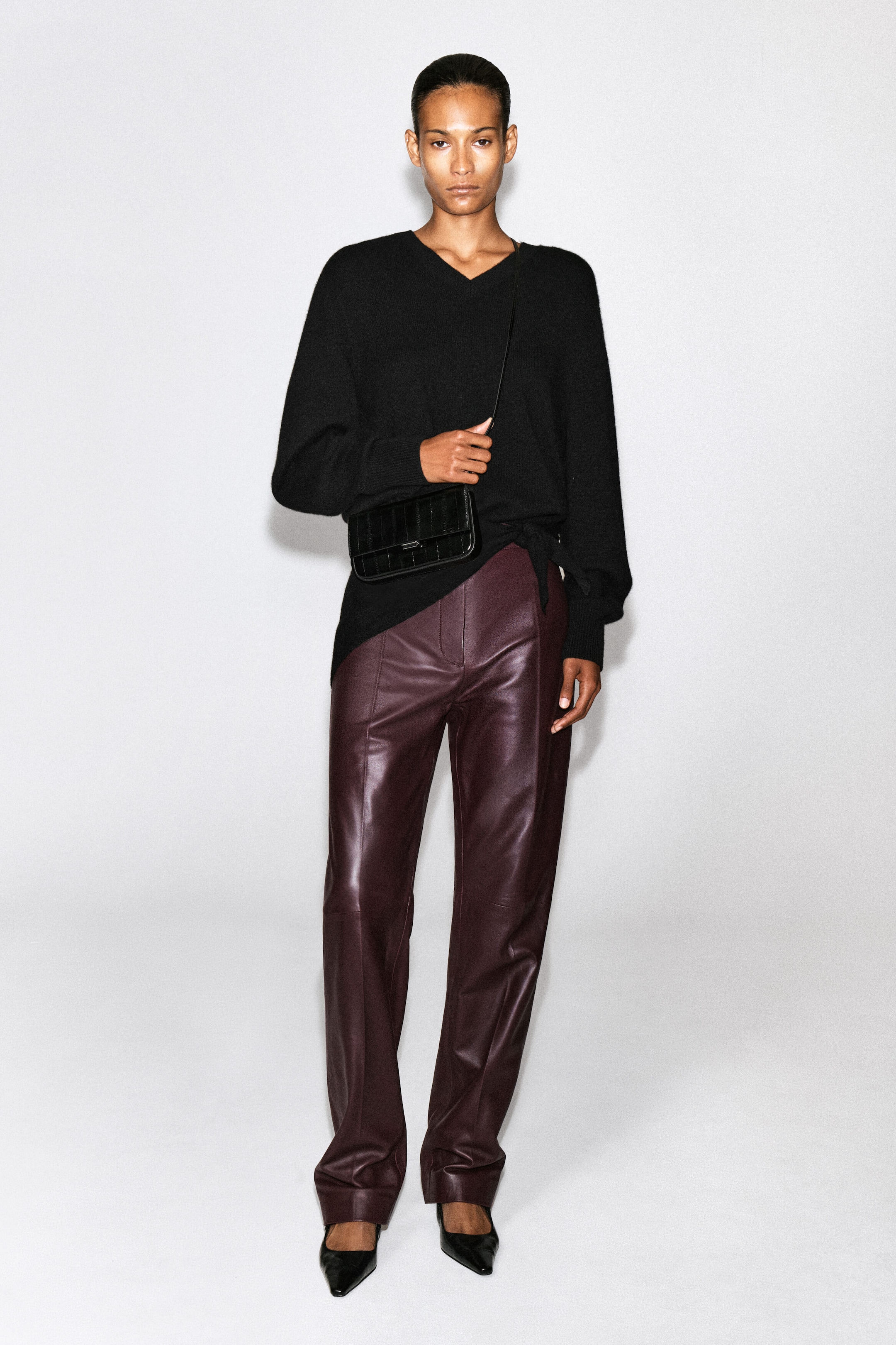 The Parquet shoulder bag in black eel-effect leather features a rectangular shape with vertical paneled stitching and a thin strap, and is worn crossbody by the model. The model is holding the bag’s strap. It is styled over a black knit sweater and paired with deep burgundy leather trousers. The Parquet shoulder bag in black eel-effect leather features a rectangular shape with vertical paneled stitching and a thin strap, and is worn crossbody by the model. The model is holding the bag’s strap. It is styled over a black knit sweater and paired with deep burgundy leather trousers.