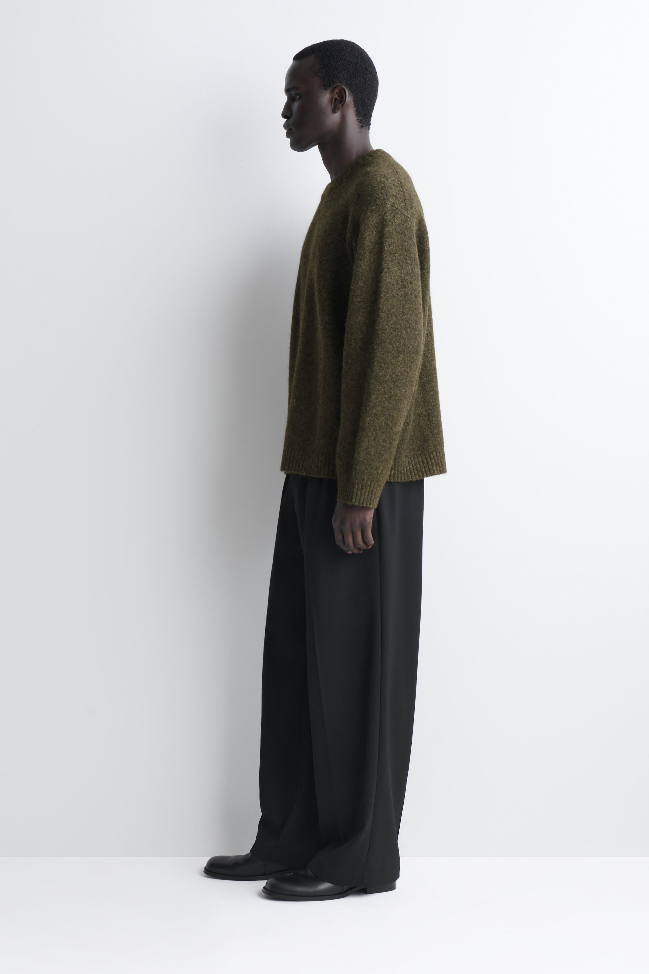 The oversized alpaca-blend crew-neck jumper in a textured olive-green knit drapes loosely on a model facing left, showcasing its relaxed silhouette and dropped shoulders. The jumper features a ribbed hem and cuffs, and is styled with wide-leg black trousers that complement its oversized fit. The oversized alpaca-blend crew-neck jumper in a textured olive-green knit drapes loosely on a model facing left, showcasing its relaxed silhouette and dropped shoulders. The jumper features a ribbed hem and cuffs, and is styled with wide-leg black trousers that complement its oversized fit.