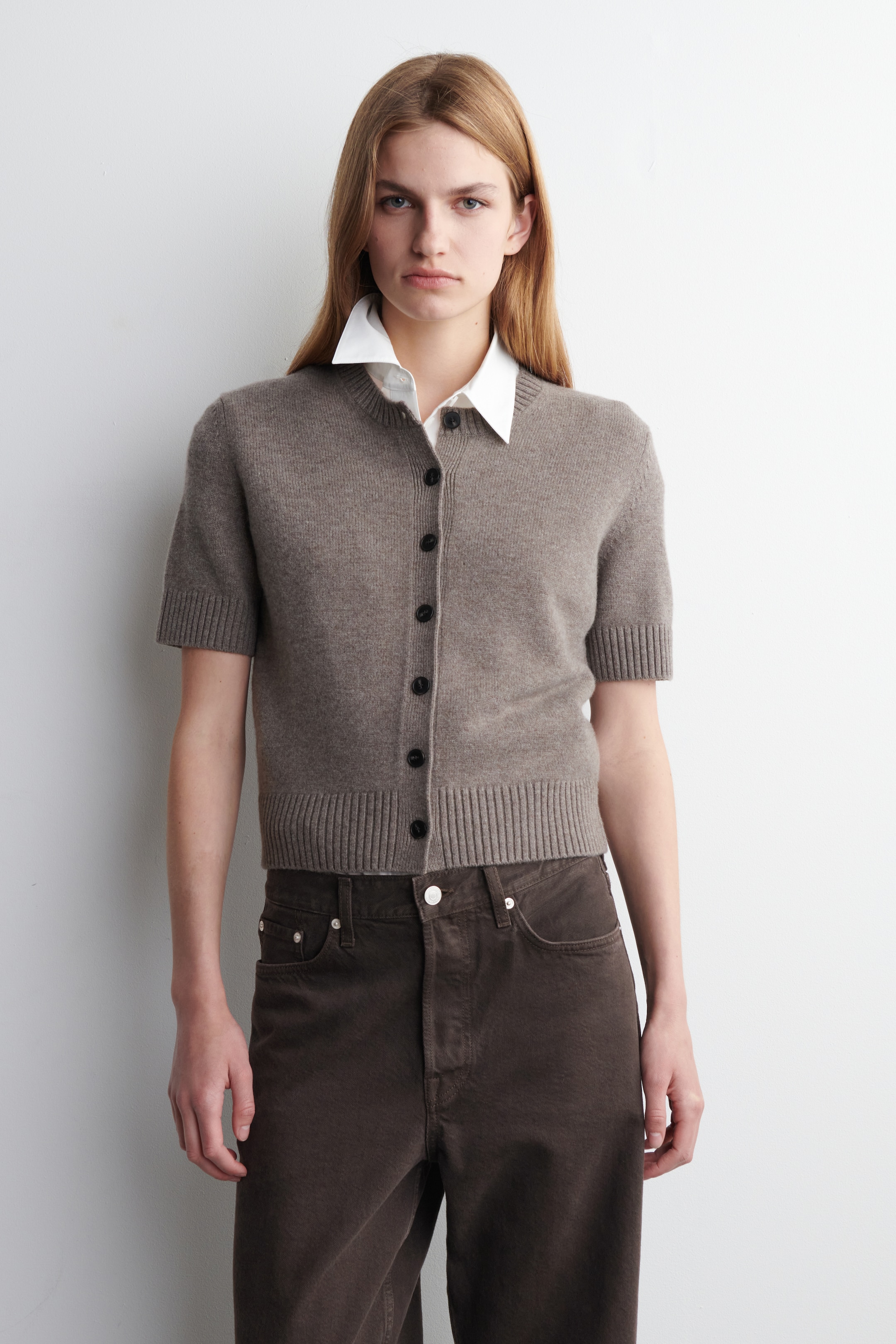 Größeres Bild ansehen: Eine taupefarbene Strickjacke mit kurzen Ärmeln wird präsentiert, die über einen Rundhalsausschnitt und eine durchgehende Knopfleiste mit dunklen, runden Knöpfen verfügt. Sie ist taillenlang geschnitten und mit einem gerippten Saum und gerippten Bündchen versehen, über einem weißen Hemd mit Kragen getragen.