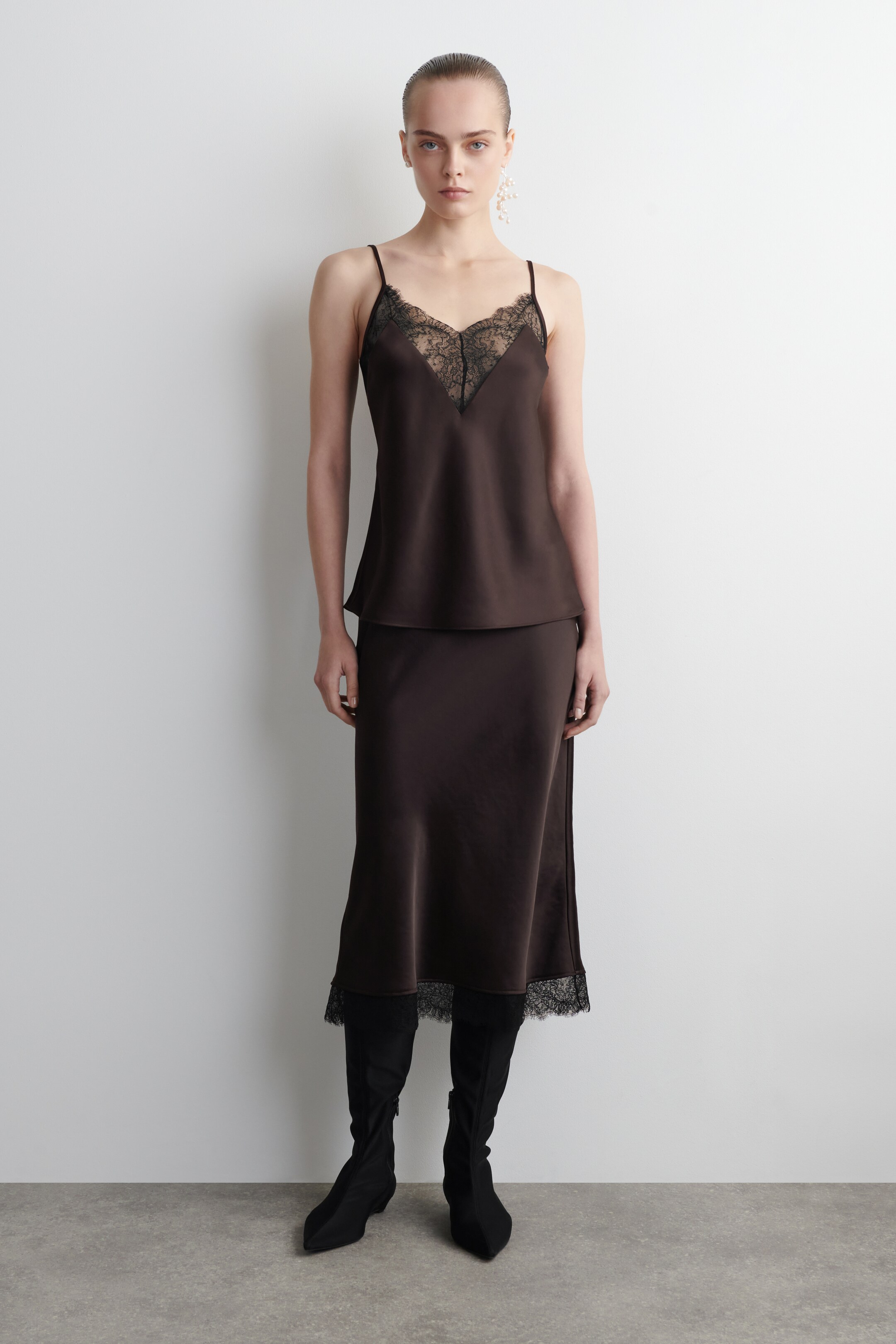 Ver imagen más grande: La camiseta de satén de tono chocolate se presenta desde el FRENTE, con un escote en V adornado con un ribete de encaje negro contrastante y finos tirantes ajustables. Está combinada con una falda a juego de tono chocolate, que también muestra detalles de encaje negro en su dobladillo.