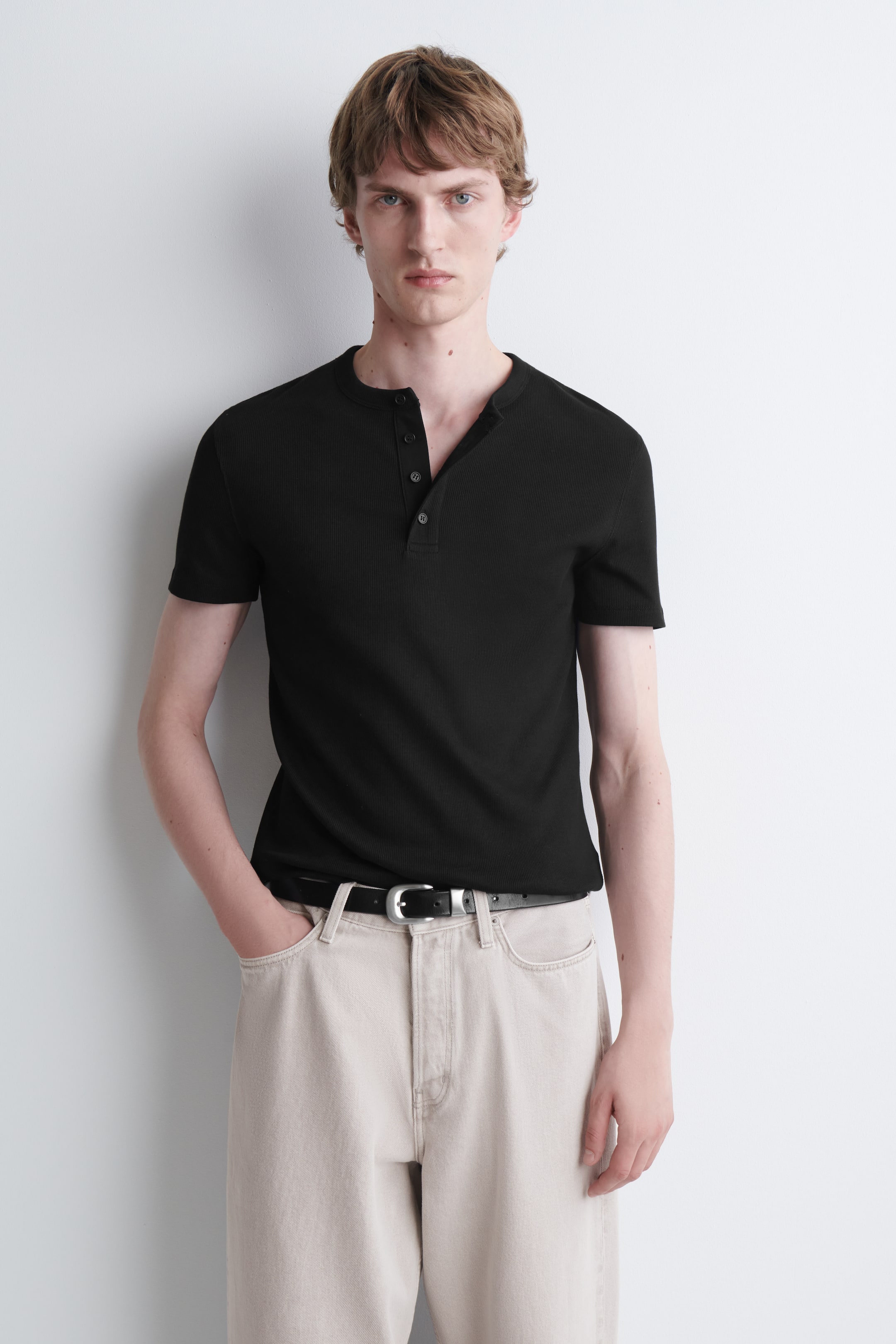 Visualizza l'immagine ingrandita: La modella indossa una maglietta henley nera, aderente e a coste, infilata in pantaloni beige chiaro con una cintura nera. La maglietta a maniche corte presenta una texture a coste e una patta con bottoni parzialmente aperta sul colletto.