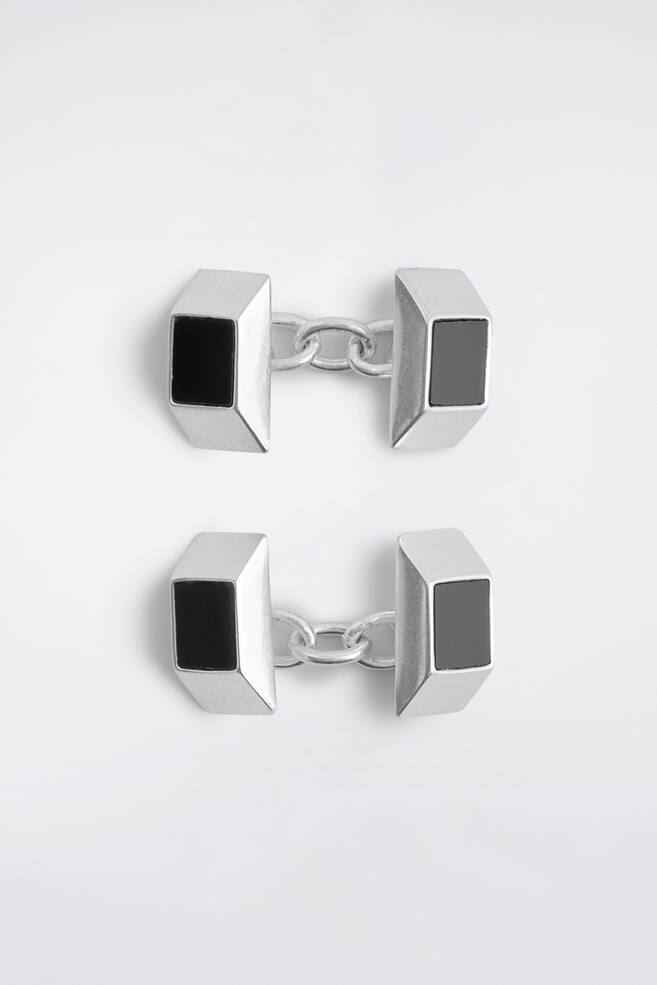 Et par manchetknapper i sÃ¸lvfarvet tone vises forfra med et facetteret, bÃ¸rstet metaldesign med indfÃ¦ldede sorte rektangulÃ¦re sten i midten. Hver manchetknap bestÃ¥r af to identiske geometriske blokke forbundet med et kort kÃ¦deled. - 1