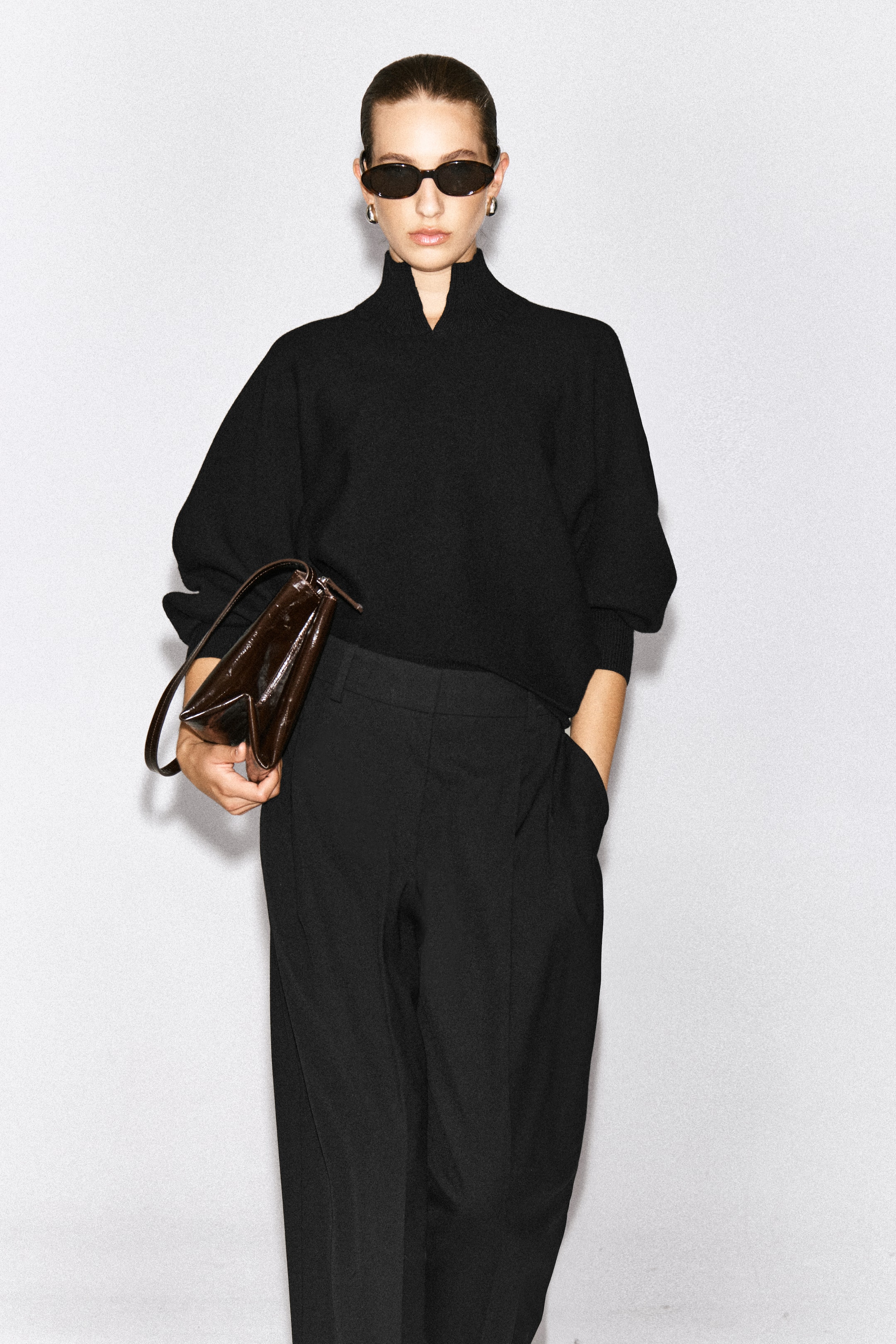 Ver imagen más grande: La modelo lleva un jersey negro de lana hervida con cuello subido tipo chimenea, un pequeño detalle en V en el delantero y puños de canalé. El jersey de corte relajado se combina con pantalones negros, mientras la modelo sostiene un bolso de piel marrón oscuro.