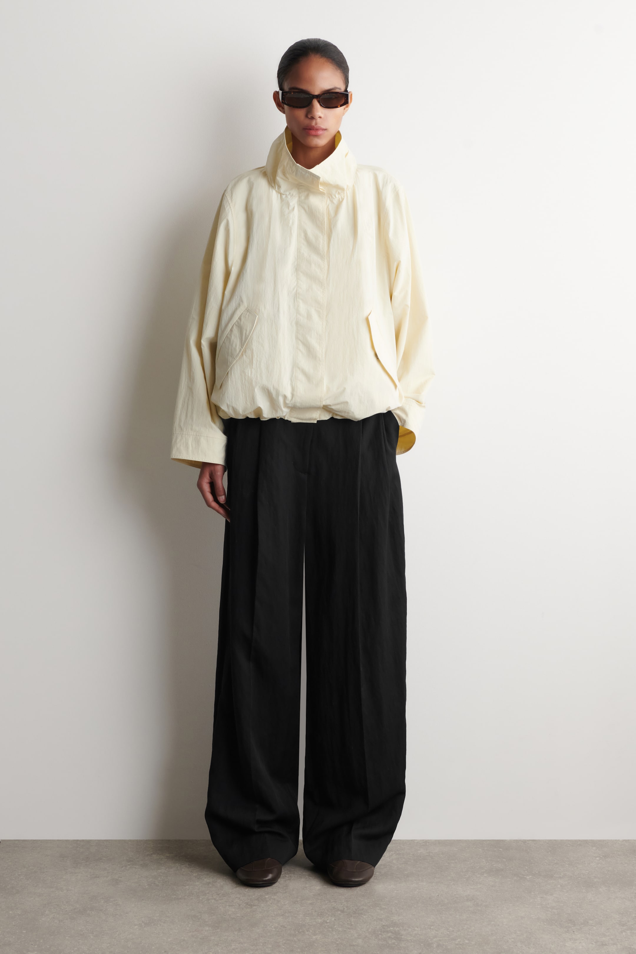 View larger image: A light yellow jacket featuring a high-standing collar and a relaxed fit. It is crafted from a lightweight, crinkled fabric and showcases a gathered hem and two front flap pockets.