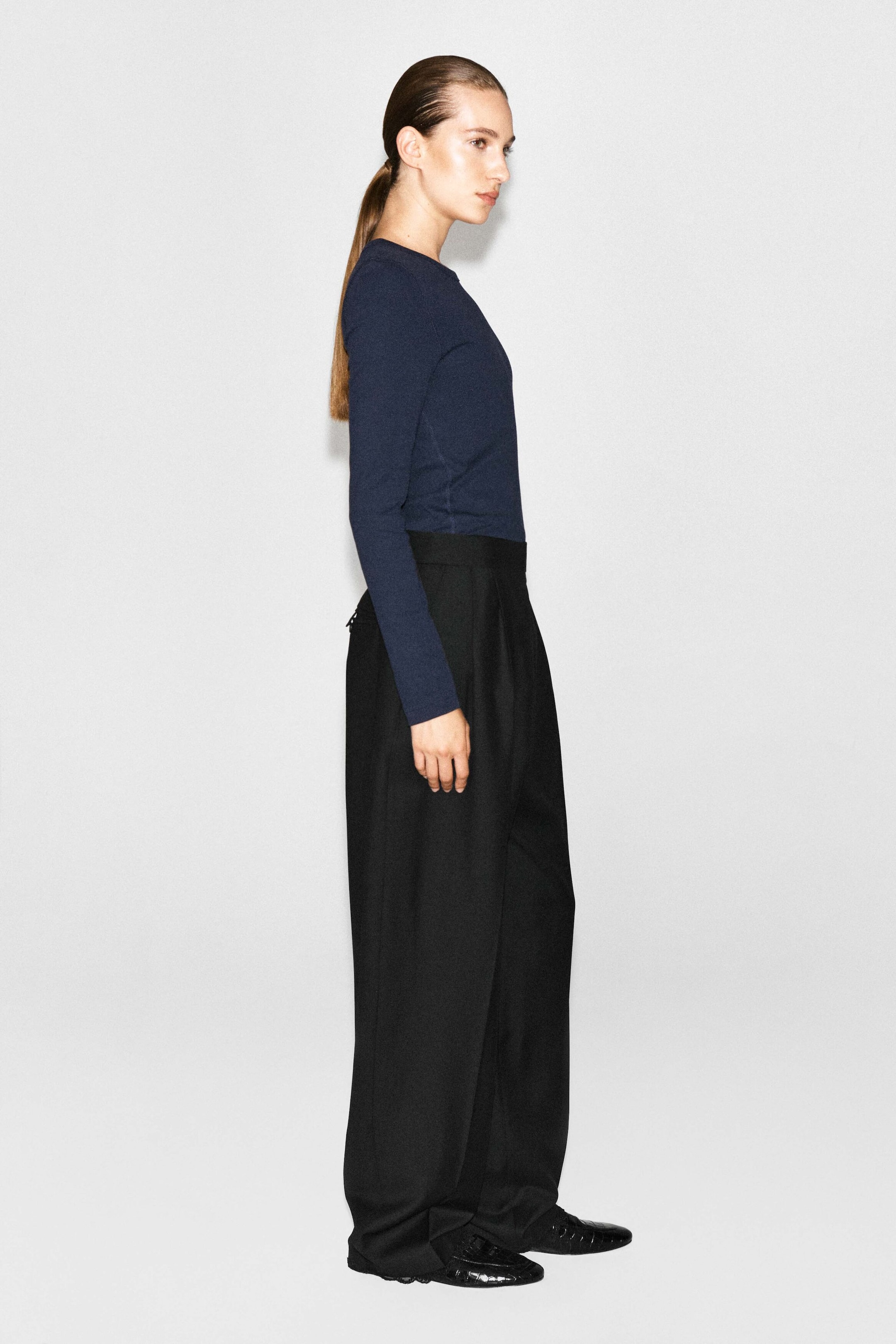 A dark blue, slim ribbed cotton long-sleeved top is being worn by the model, tucked into high-waisted, wide-leg black trousers. The top features a crew neckline and a close-fitting silhouette along the arms and torso. A dark blue, slim ribbed cotton long-sleeved top is being worn by the model, tucked into high-waisted, wide-leg black trousers. The top features a crew neckline and a close-fitting silhouette along the arms and torso.