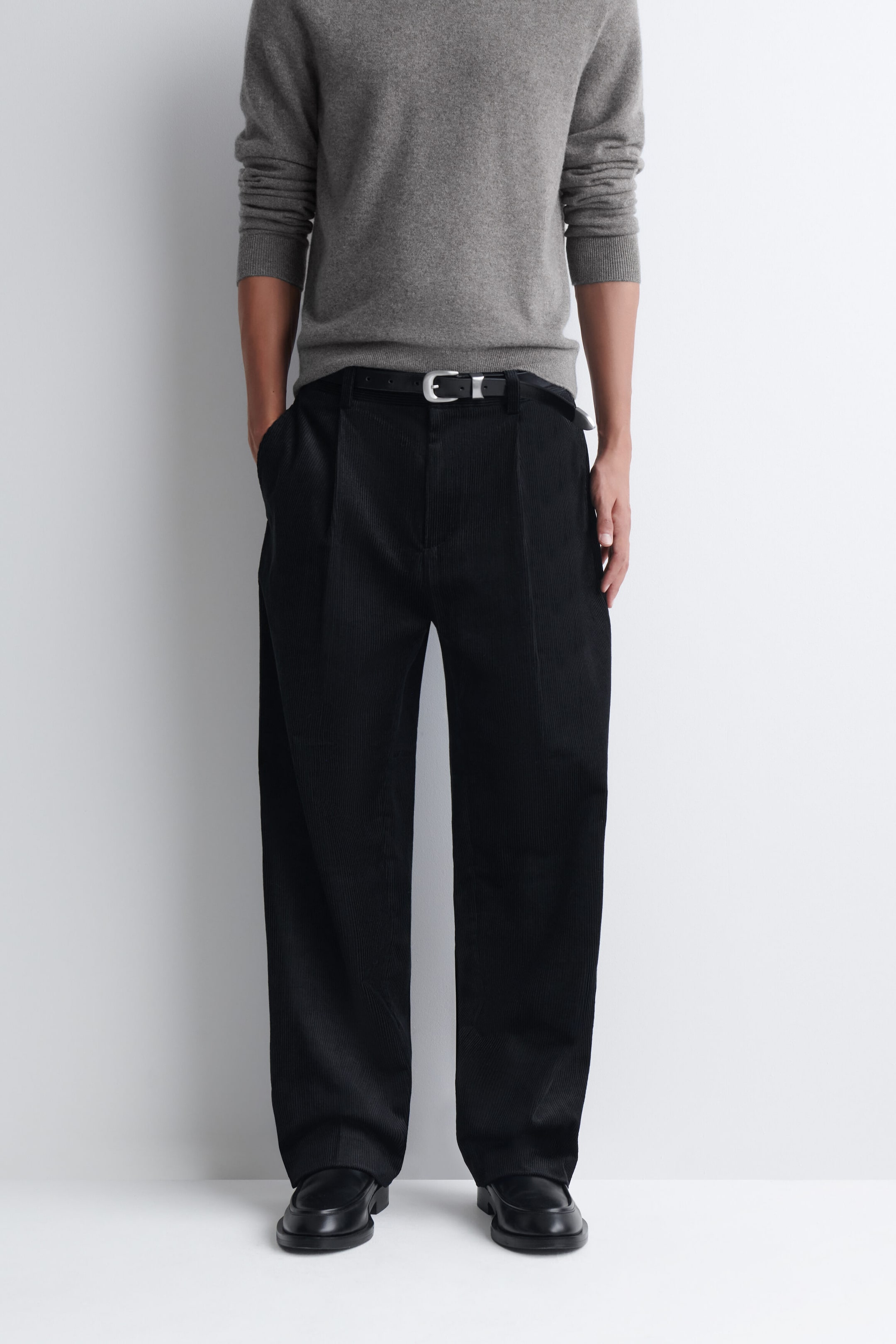 Pleated relaxed-fit tapered corduroy pants in black are worn by the model, who is standing with one hand resting in a side pocket. The pants feature front pleats and are styled with a black leather belt, a grey long-sleeved sweater, and black leather shoes. Pleated relaxed-fit tapered corduroy pants in black are worn by the model, who is standing with one hand resting in a side pocket. The pants feature front pleats and are styled with a black leather belt, a grey long-sleeved sweater, and black leather shoes.