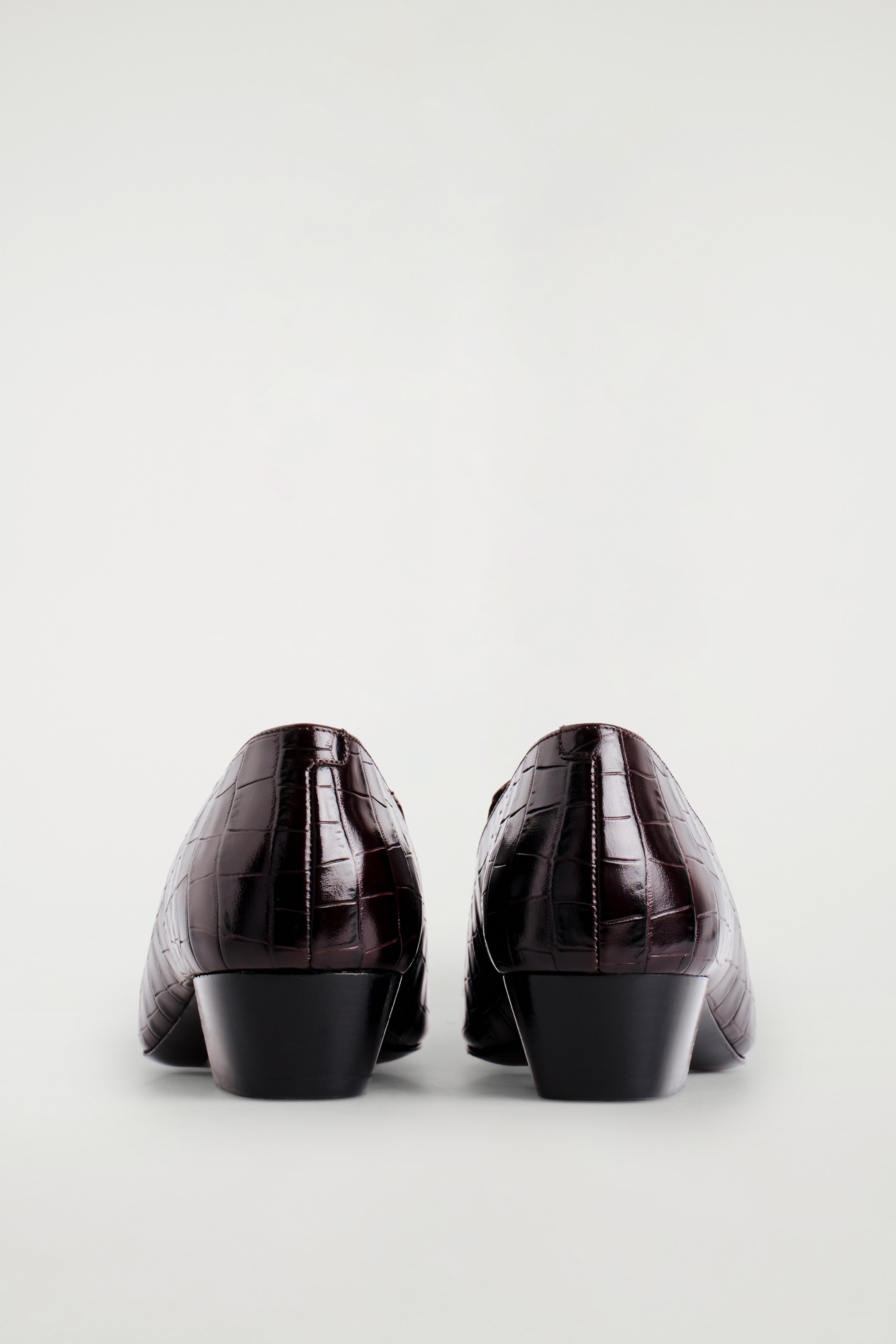 Agrandir l'image: Une paire de mocassins marron foncé et brillants est montrée de dos. Ils présentent un motif embossé à effet crocodile et des talons blocs angulaires de 4 cm.