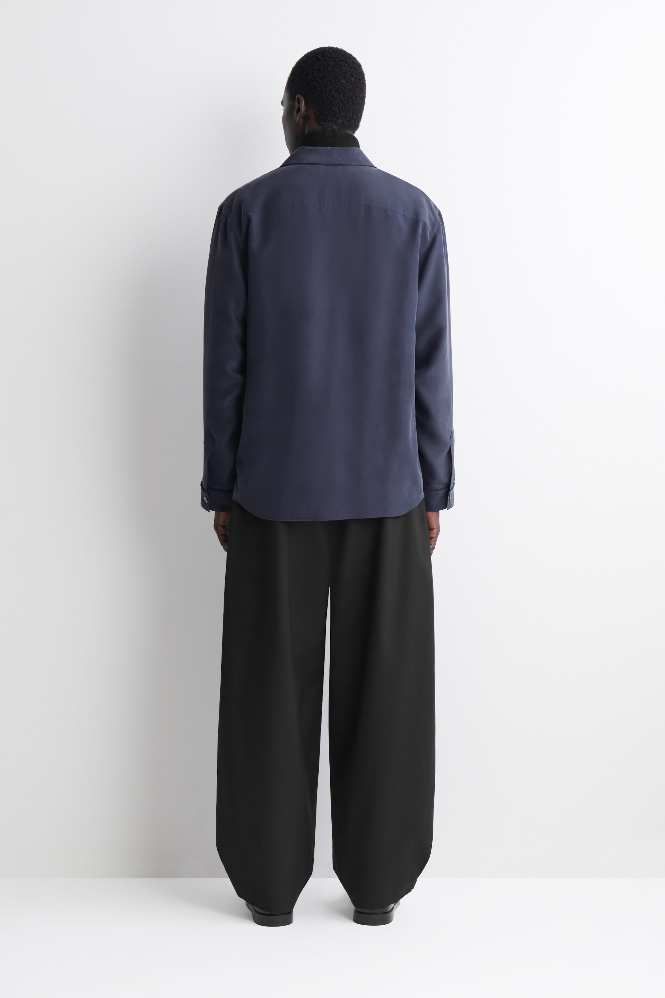 A long-sleeved navy blue silk shirt is shown from the back, highlighting its relaxed fit, straight back yoke, and soft shawl collar. The shirt is layered over a black high-neck top and paired with wide-leg black trousers to showcase its fluid drape. A long-sleeved navy blue silk shirt is shown from the back, highlighting its relaxed fit, straight back yoke, and soft shawl collar. The shirt is layered over a black high-neck top and paired with wide-leg black trousers to showcase its fluid drape.