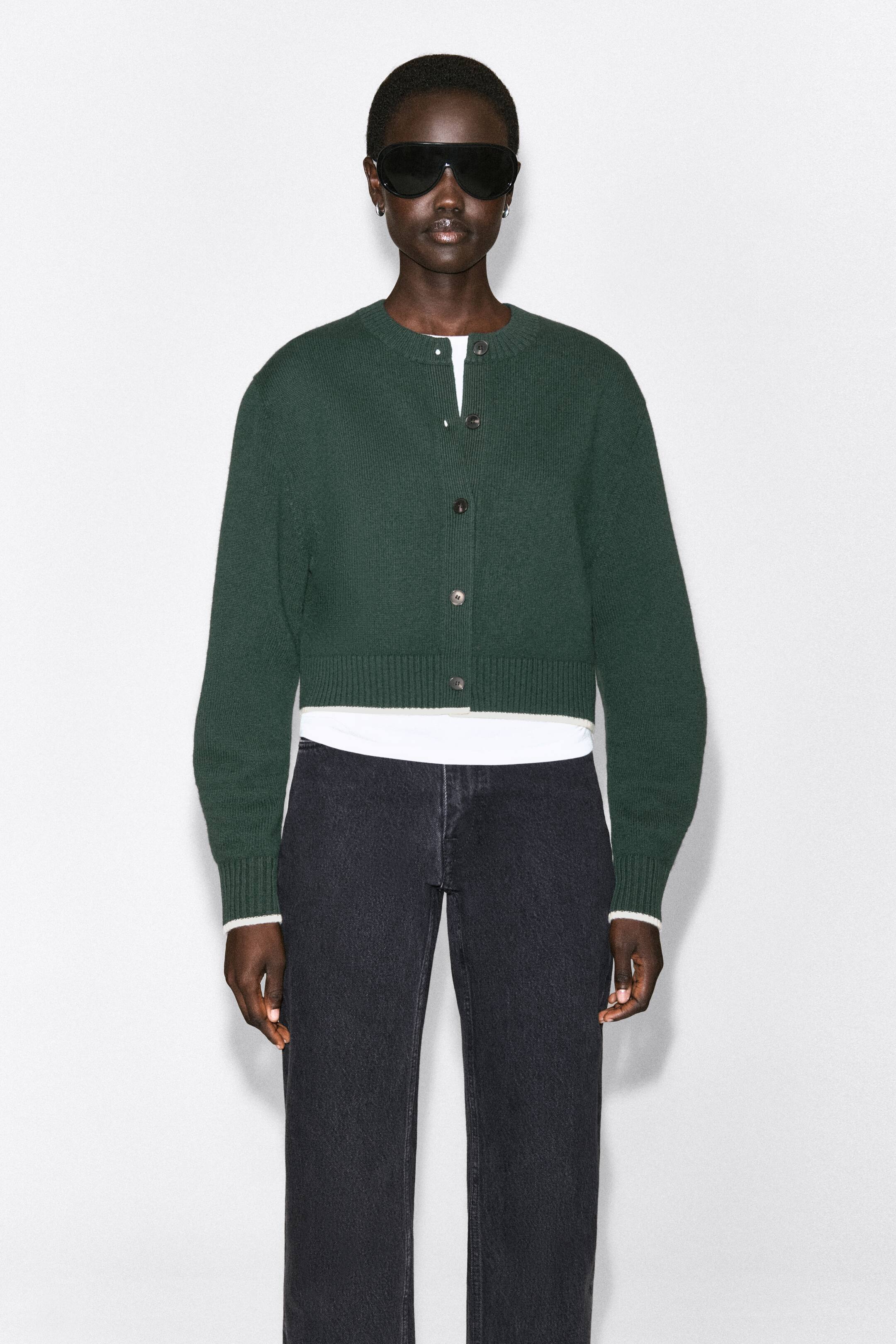 Ver imagen más grande: Esta cárdigan de lana merina verde oscuro está vista de frente, con una silueta cropped, cuello redondo, cierre frontal con botones y puños y bajo de punto acanalado. Se lleva superpuesta sobre una camiseta blanca que asoma en el cuello, los puños y el bajo, destacando su corte estructurado y cropped.