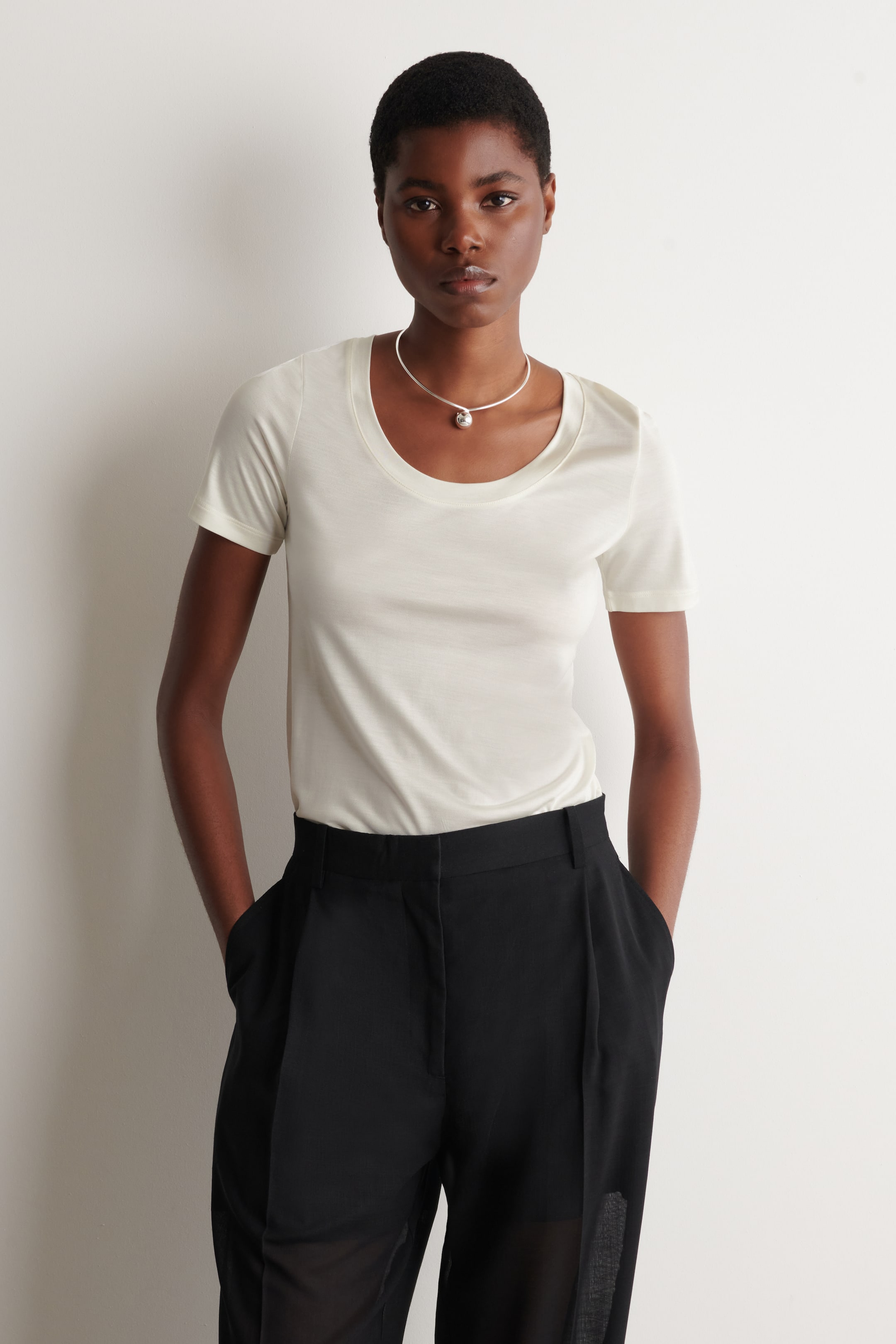 Se større billede: En dæmpet elfenbensfarvet T-shirt med korte ærmer og en rund halsudskæring bæres af modellen. Den er stoppet ned i sorte bukser, og en sølv choker-halskæde hviler på halsudskæringen.