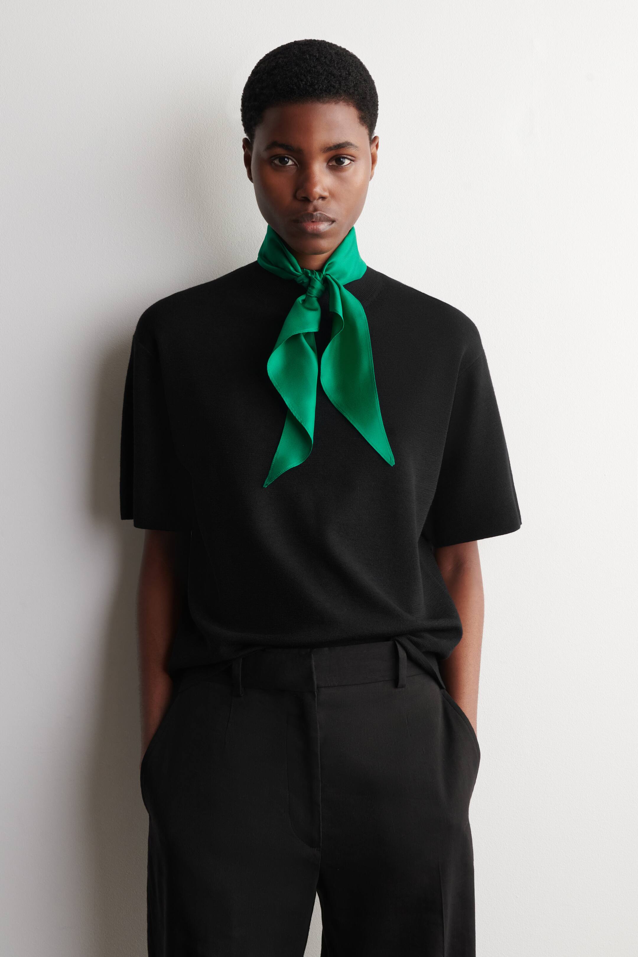 Agrandir l'image: Un foulard vert vif et brillant est noué à l'avant du cou, ses extrémités pointues s'évasant sur un haut noir à manches courtes.