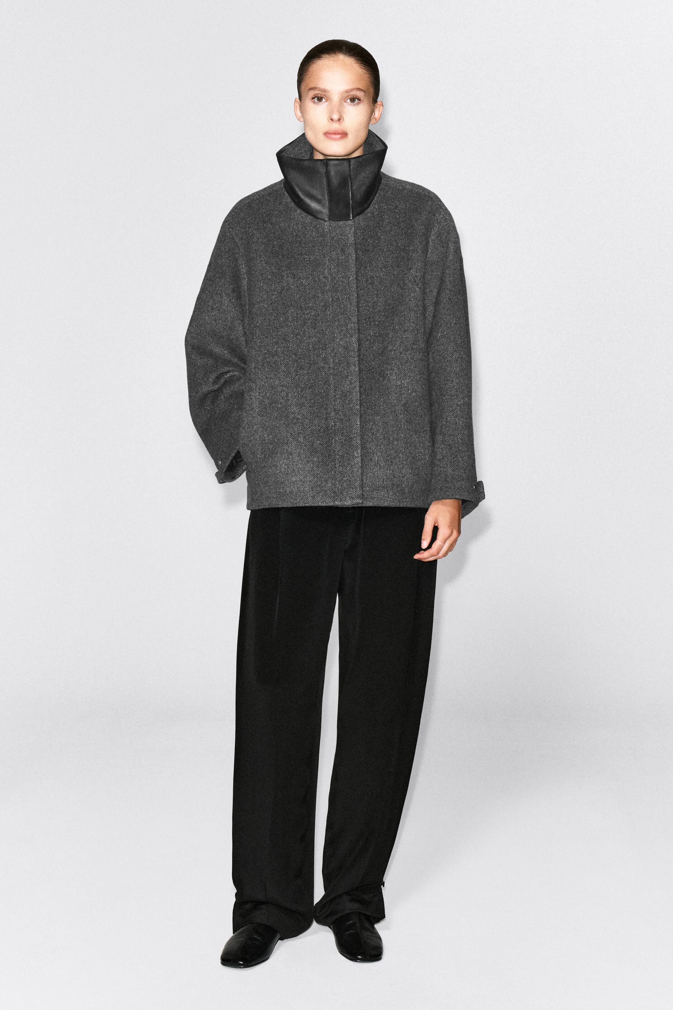 Se større billede: En grå jakke i fiskebensmønster med høj, struktureret sort læderfunnelhals og oversized silhuet bæres af modellen. Jakken styles med sorte bukser med brede ben, mens modellen står med den ene hånd placeret i sidelommen.