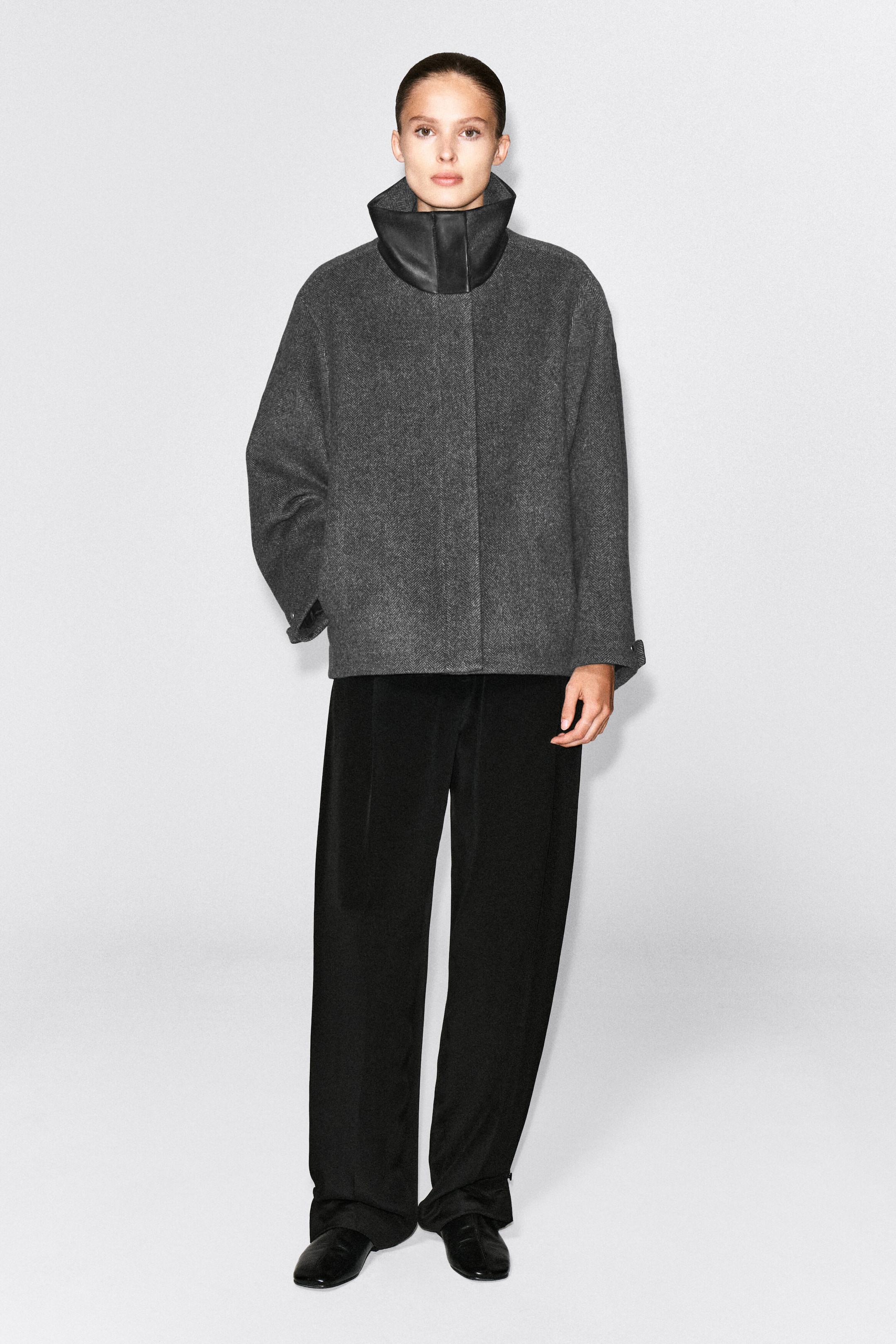 Ein grauer Fischgrat-Blazer mit hohem, strukturiertem schwarzem Leder-Trichterausschnitt und Oversize-Silhouette wird vom Model getragen. Der Blazer ist mit weit geschnittenen schwarzen Hosen kombiniert, wobei das Model steht und eine Hand in der Seitentasche hat. Ein grauer Fischgrat-Blazer mit hohem, strukturiertem schwarzem Leder-Trichterausschnitt und Oversize-Silhouette wird vom Model getragen. Der Blazer ist mit weit geschnittenen schwarzen Hosen kombiniert, wobei das Model steht und eine Hand in der Seitentasche hat.