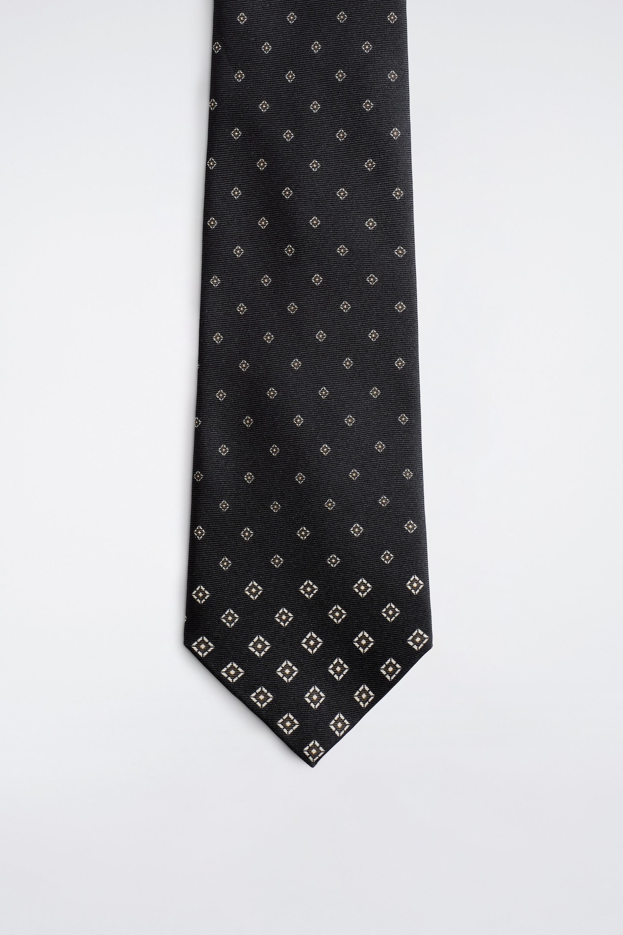 A black silk twill tie is shown from the front, featuring a repeating foulard pattern of small geometric motifs that gradually increase in size toward the pointed tip.