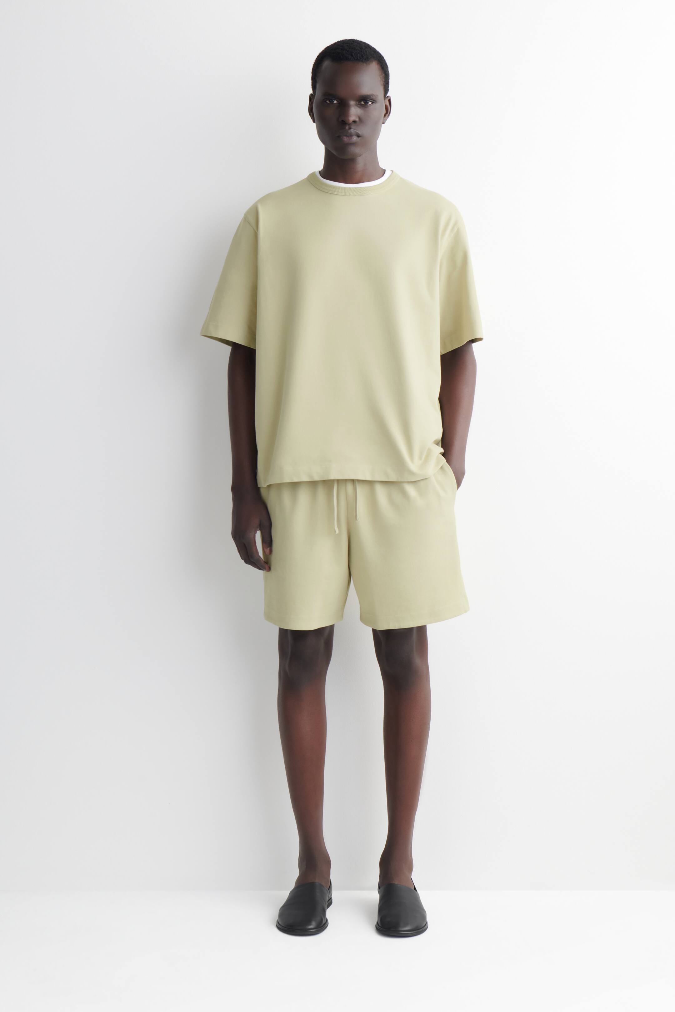 Visualizza immagine più grande: Un set composto da una maglietta a maniche corte di colore verde-giallo chiaro e pantaloncini coordinati presenta una vestibilità rilassata, rivolto in avanti. La maglietta è sovrapposta a una t-shirt bianca con scollo a girocollo, e i pantaloncini sono progettati con una fascia in vita con coulisse.