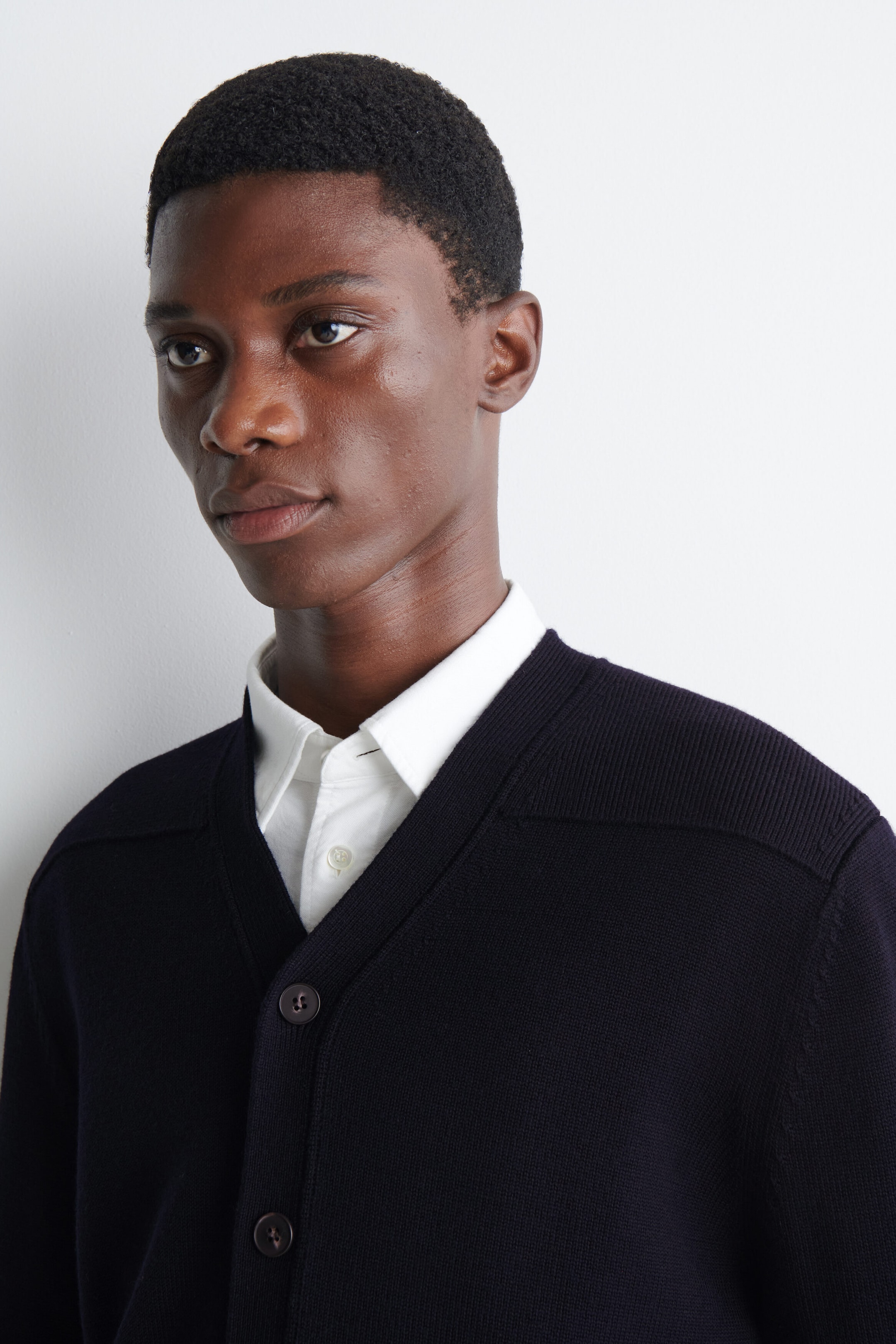 A dark navy knitted cardigan with a V-neck and button fastenings is being worn by the model. The cardigan is layered over a white, button-up collared shirt. A dark navy knitted cardigan with a V-neck and button fastenings is being worn by the model. The cardigan is layered over a white, button-up collared shirt.
