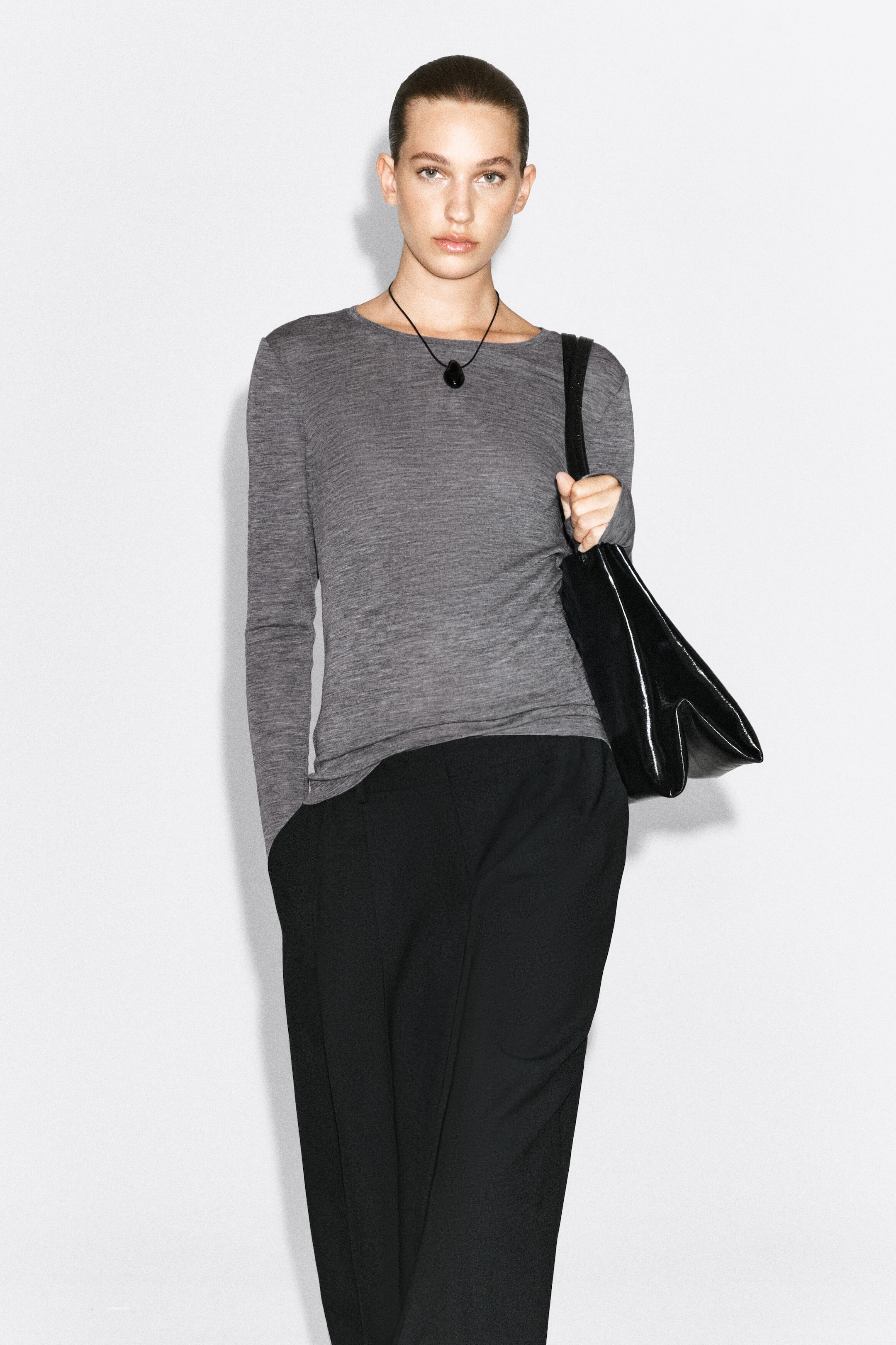 A slim-fit, long-sleeved grey merino wool top featuring a wide round neckline is being worn by the model. The top is paired with black trousers, a black pendant necklace, and a black shoulder bag carried on the shoulder.