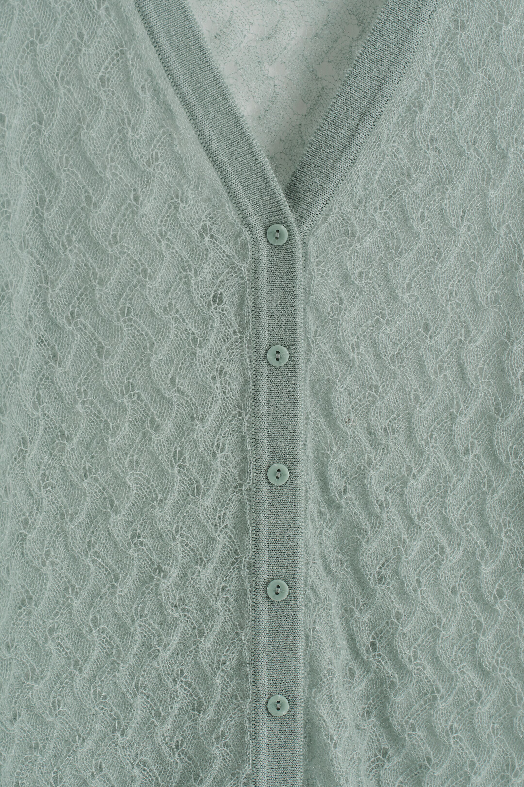 Größeres Bild anzeigen: Die Vorderseite einer hellgrünen V-Ausschnitt-Strickjacke wird gezeigt, die ein zartes Pointelle-Strickmuster aufweist, das ein subtil durchsichtiges Finish erzeugt. Eine Reihe passender Knöpfe verläuft entlang der vorderen Mitte, eingerahmt von gerippten Blenden.