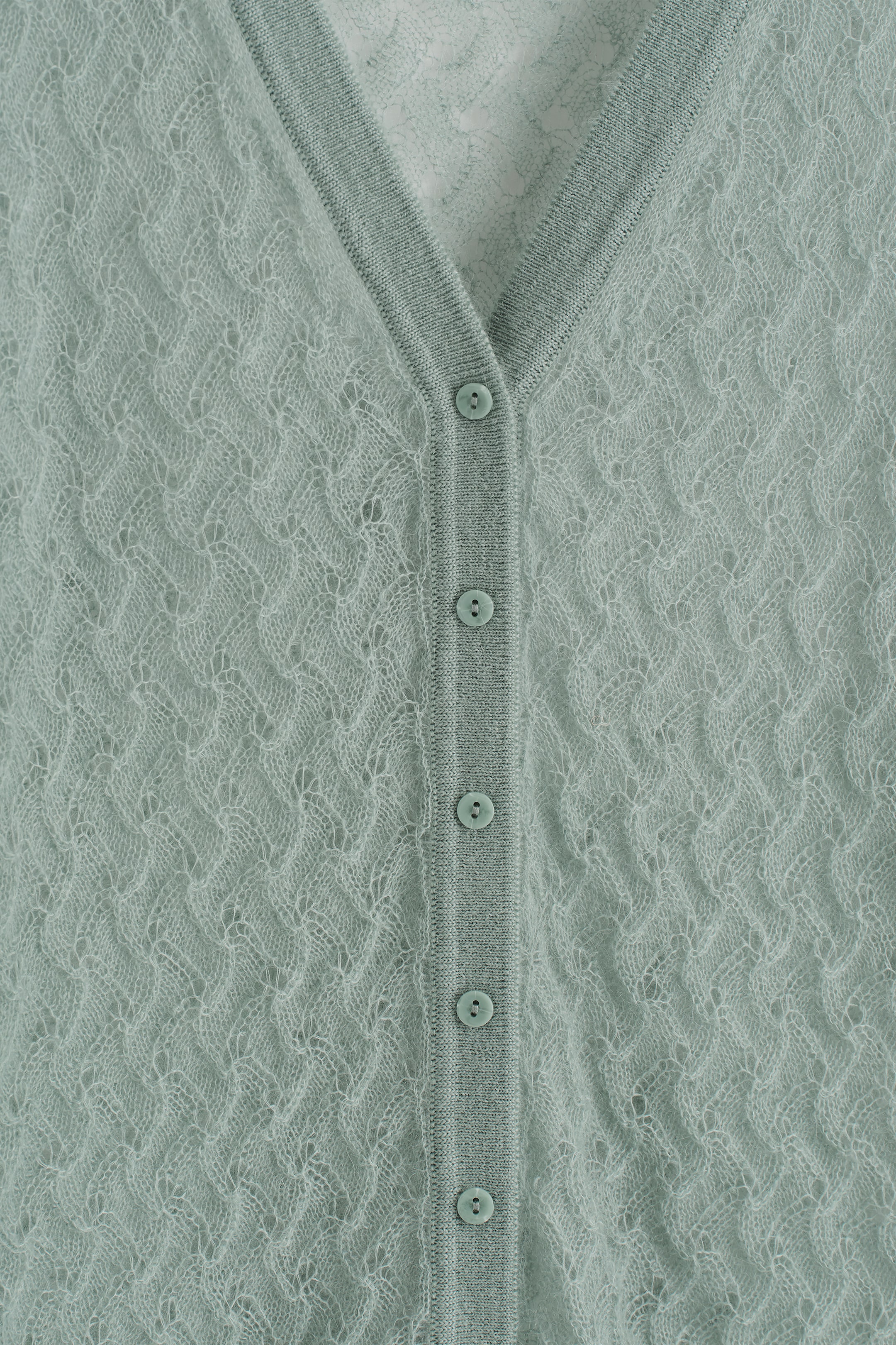 Grösseres Bild ansehen: Die Vorderseite einer hellgrünen V-Ausschnitt-Strickjacke wird gezeigt, die ein zartes Pointelle-Strickmuster aufweist, das ein subtil durchsichtiges Finish erzeugt. Eine Reihe passender Knöpfe verläuft entlang der vorderen Mitte, eingerahmt von gerippten Blenden.