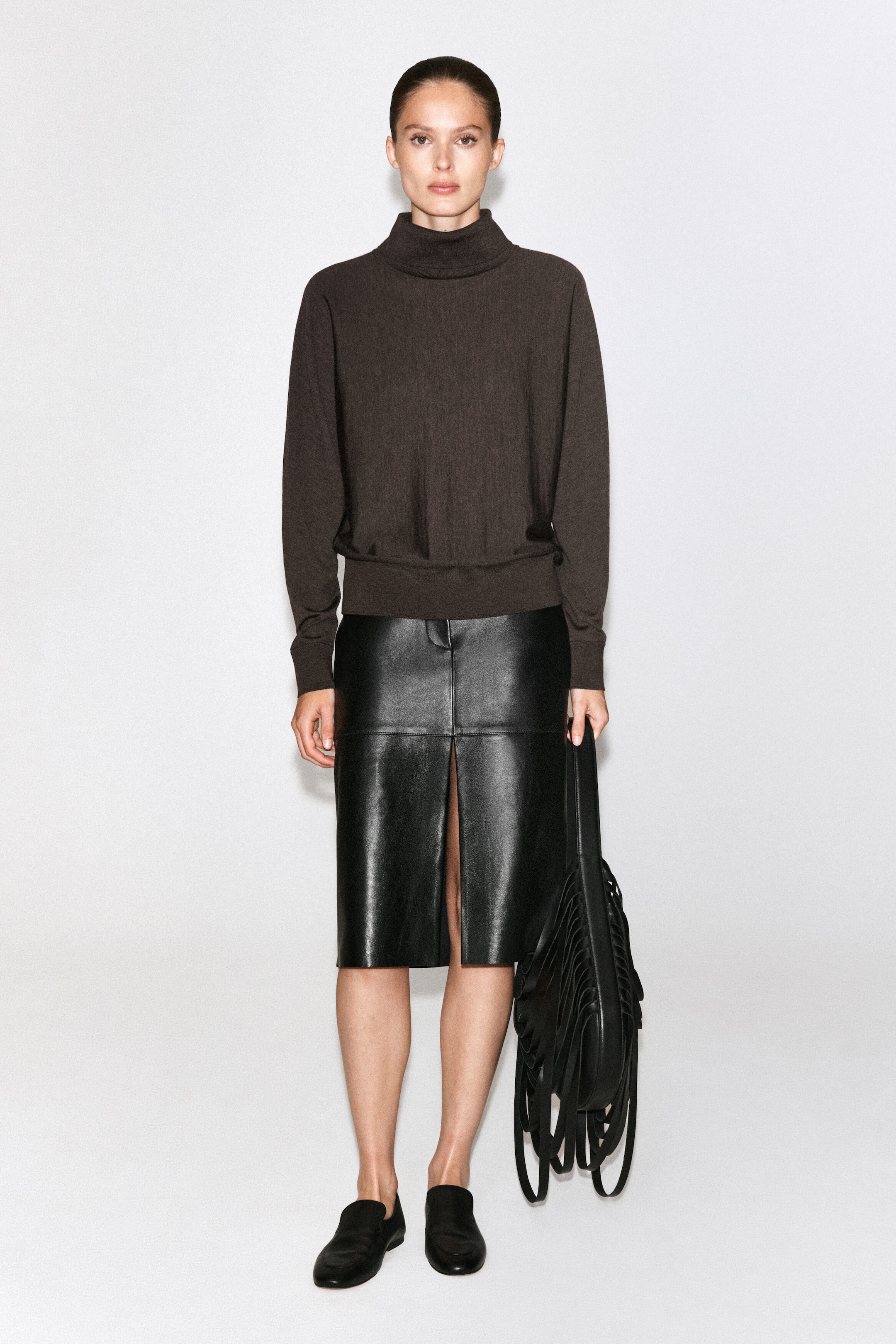The model is wearing a dark brown knit roll-neck jumper and a black leather knee-length skirt with a front slit. She is also carrying a black leather shoulder bag with long fringe detailing. The model is wearing a dark brown knit roll-neck jumper and a black leather knee-length skirt with a front slit. She is also carrying a black leather shoulder bag with long fringe detailing.