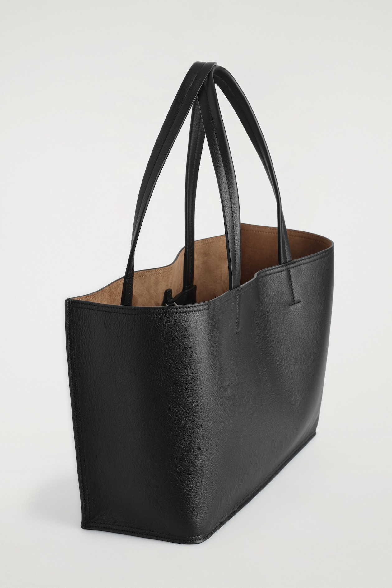A black pebbled leather tote bag is positioned at an angle facing right, showcasing its structured rectangular shape and dual flat shoulder straps. The open top reveals a contrasting tan, suede-like interior lining.