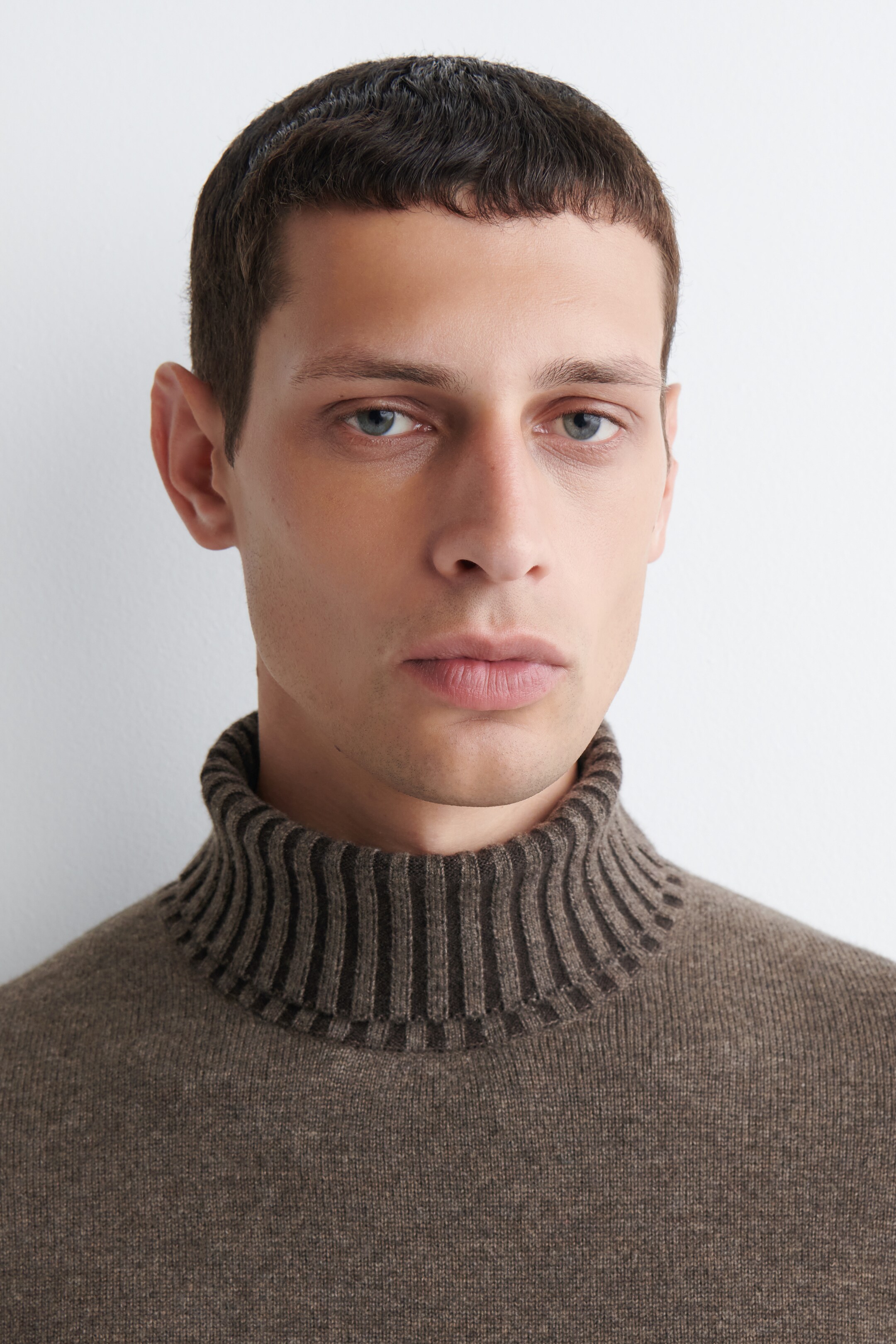 View larger image: A light brown knit turtleneck sweater is being presented, with its vertically ribbed collar prominently displayed from a front-facing view. The collar features dark brown horizontal stripes, contrasting with the smooth, light brown knit of the sweater's body.