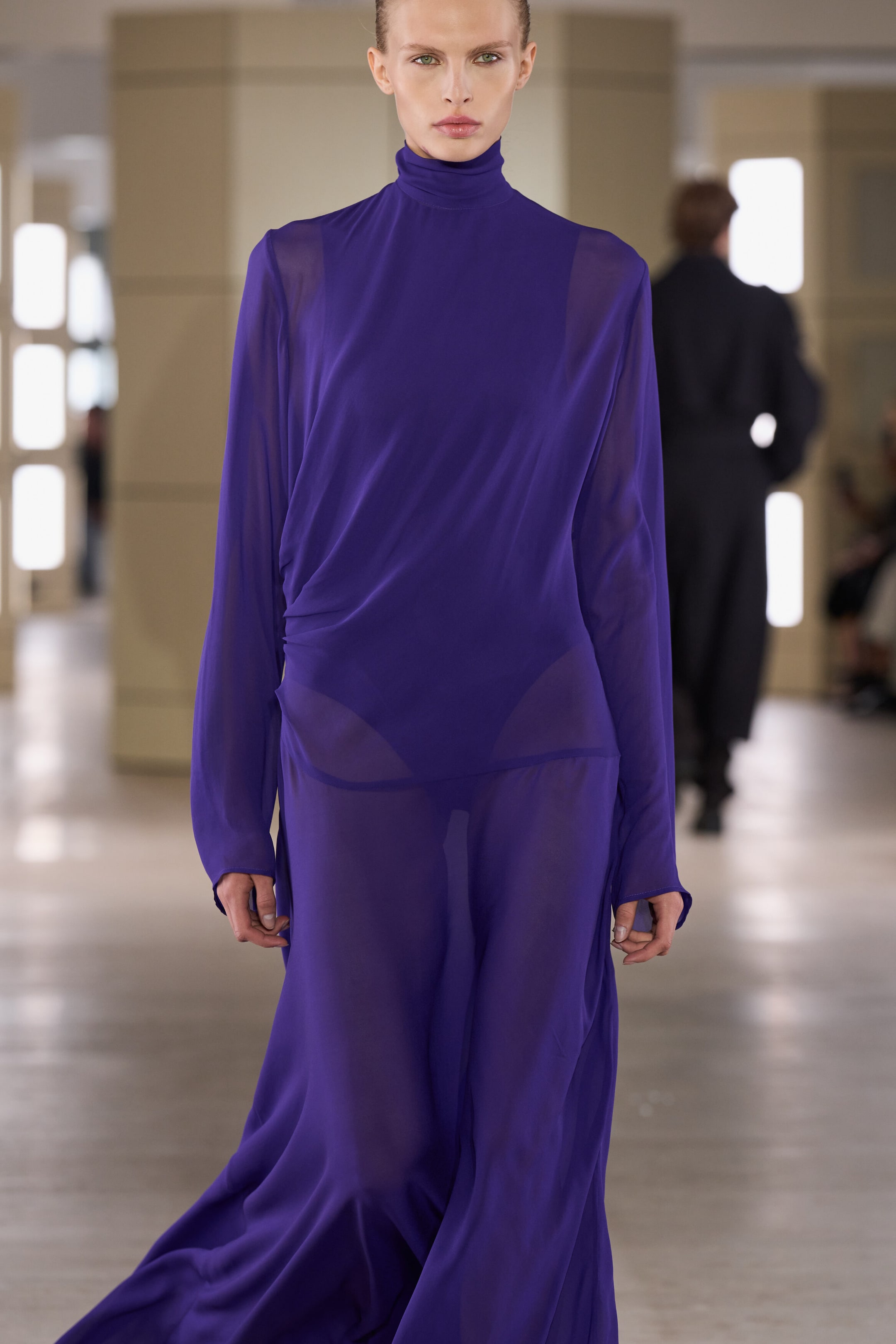 A deep purple draped high-neck maxi dress is shown from the front, featuring a high mock neck, long sleeves and sheer, flowing fabric. The dress drapes diagonally across the torso and gathers at the waist, revealing a matching purple undergarment beneath the semi-transparent skirt.