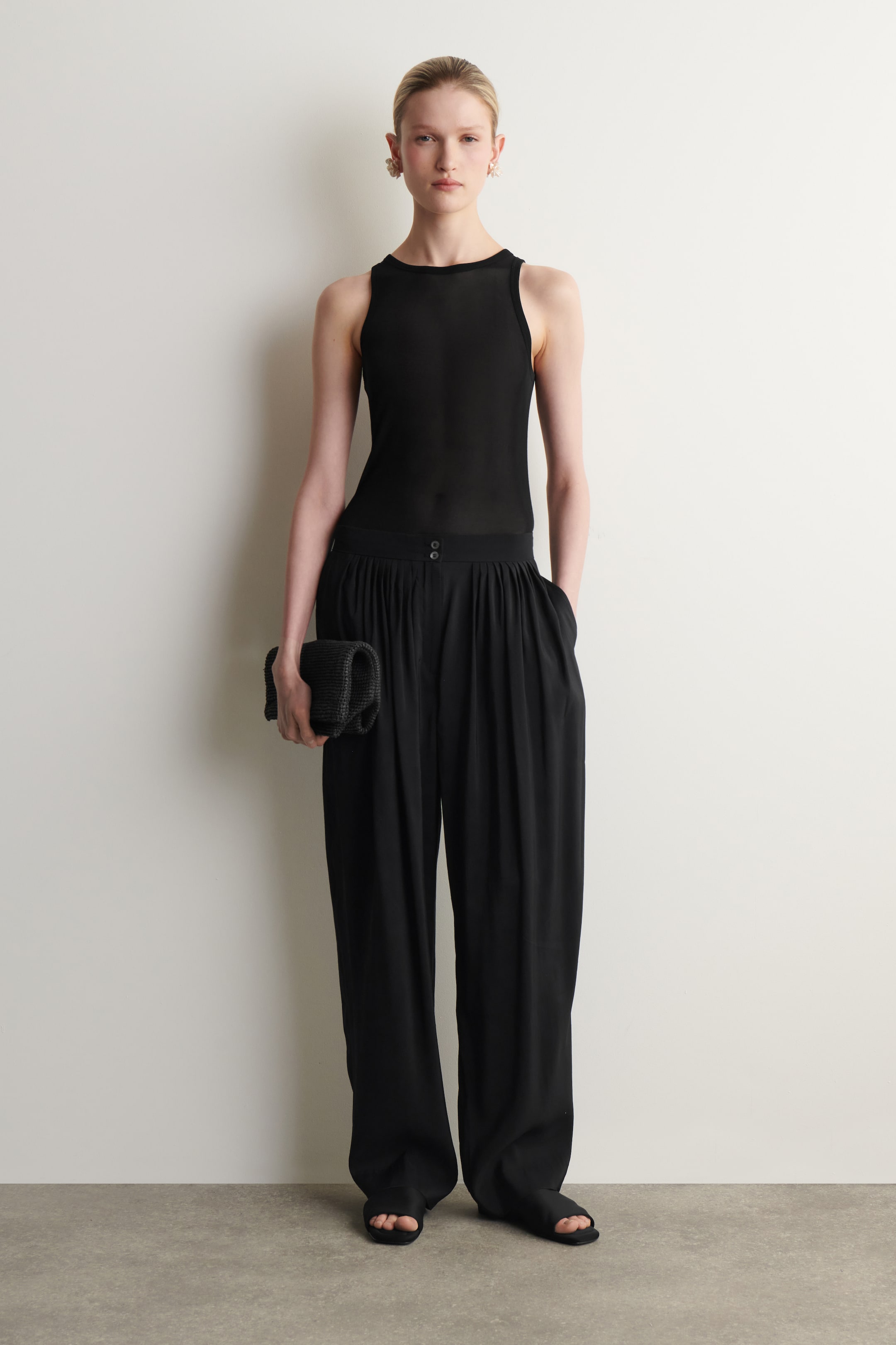 הצגת תמונה גדולה יותר: חולצה שחורה ללא שרוולים נלבשת עם מכנסיים שחורים בעלי מותן גבוהה ורגל רחבה, הכוללים קפלים ושני כפתורים בקו המותן. התלבושת מעוצבת עם קלאץ' שחור במרקם וסנדלים שחורים עם אצבעות פתוחות.