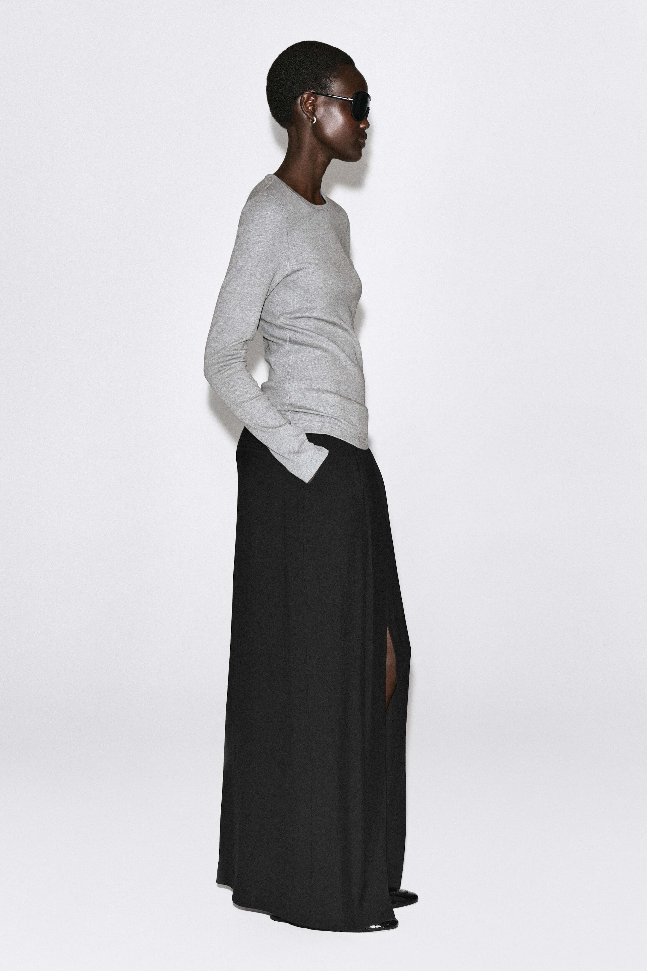 The slim ribbed cotton long-sleeve top in light grey is shown from a right-facing side profile, highlighting its close fit, crew neckline, and fine ribbed texture. It is styled over a long black maxi skirt, showing how the clean hem drapes smoothly at the hip. The slim ribbed cotton long-sleeve top in light grey is shown from a right-facing side profile, highlighting its close fit, crew neckline, and fine ribbed texture. It is styled over a long black maxi skirt, showing how the clean hem drapes smoothly at the hip.