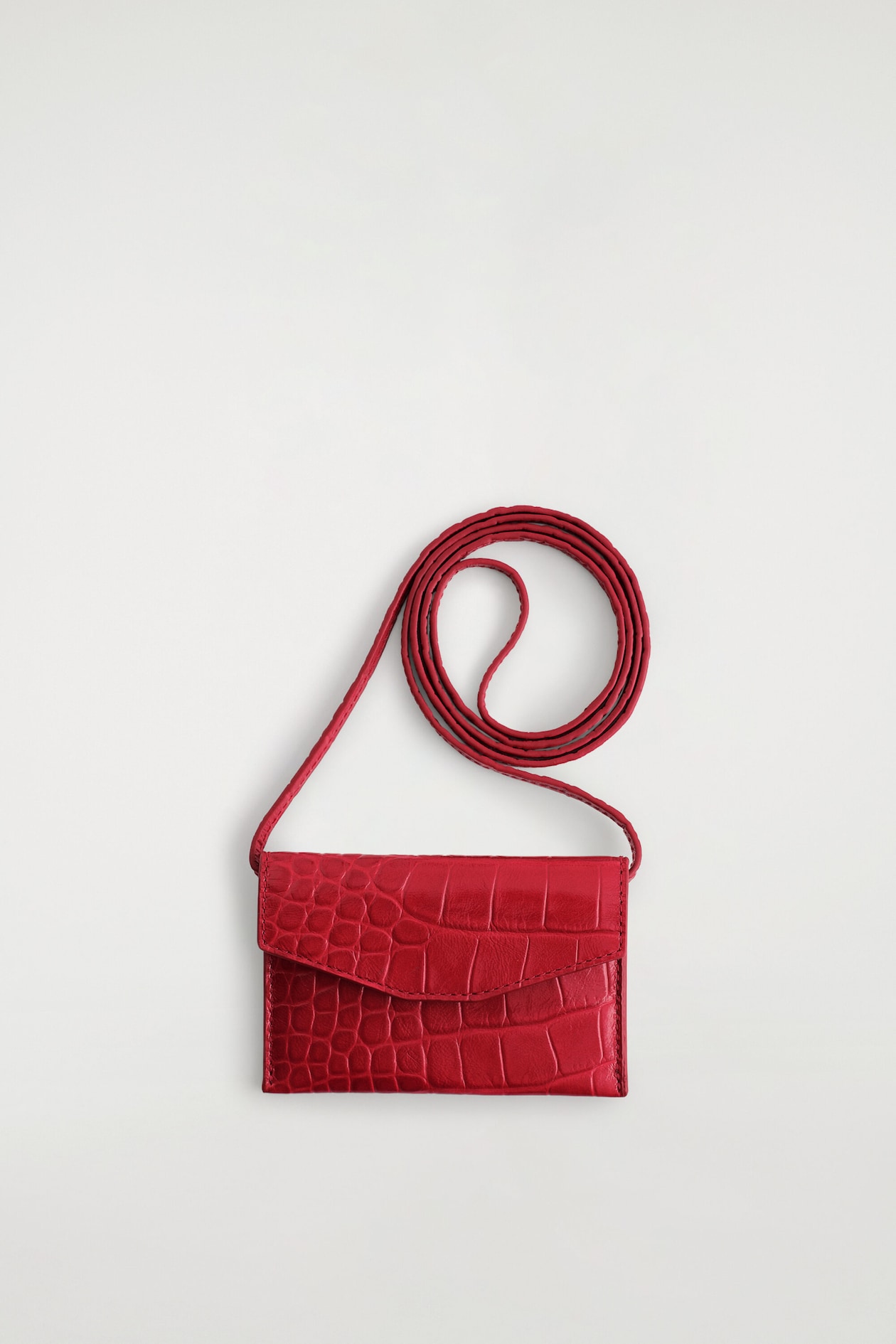 A red crocodile-effect leather card holder lies flat, facing forward to display its envelope-style flap and textured finish. A matching long strap extends from the sides and coils neatly above the card holder.