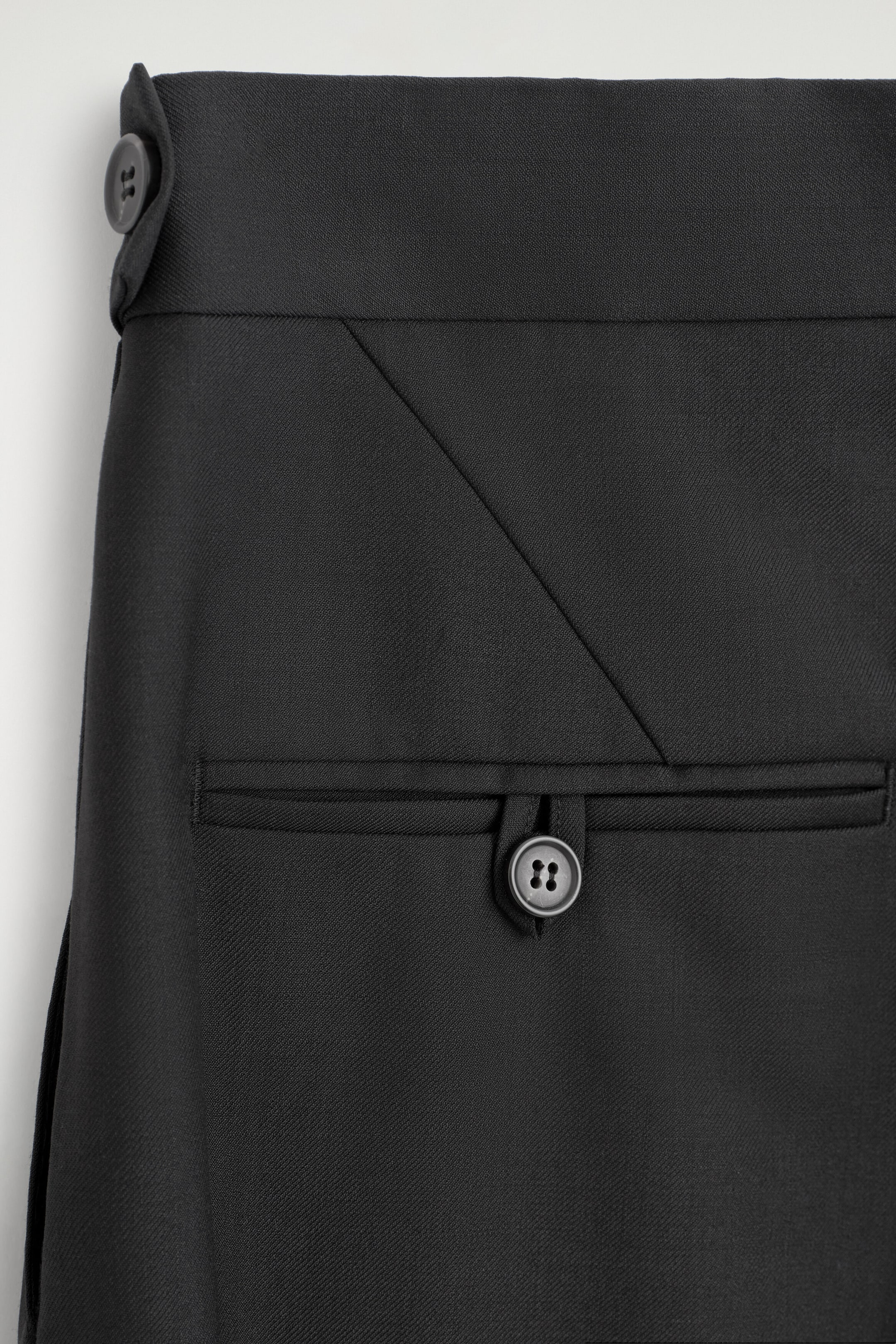 A close-up of black trousers showcasing the waist and pocket details, featuring a horizontal welt pocket with a single button-loop closure. The waistband displays an adjustable side tab with a button and a diagonal seam detail. A close-up of black trousers showcasing the waist and pocket details, featuring a horizontal welt pocket with a single button-loop closure. The waistband displays an adjustable side tab with a button and a diagonal seam detail.