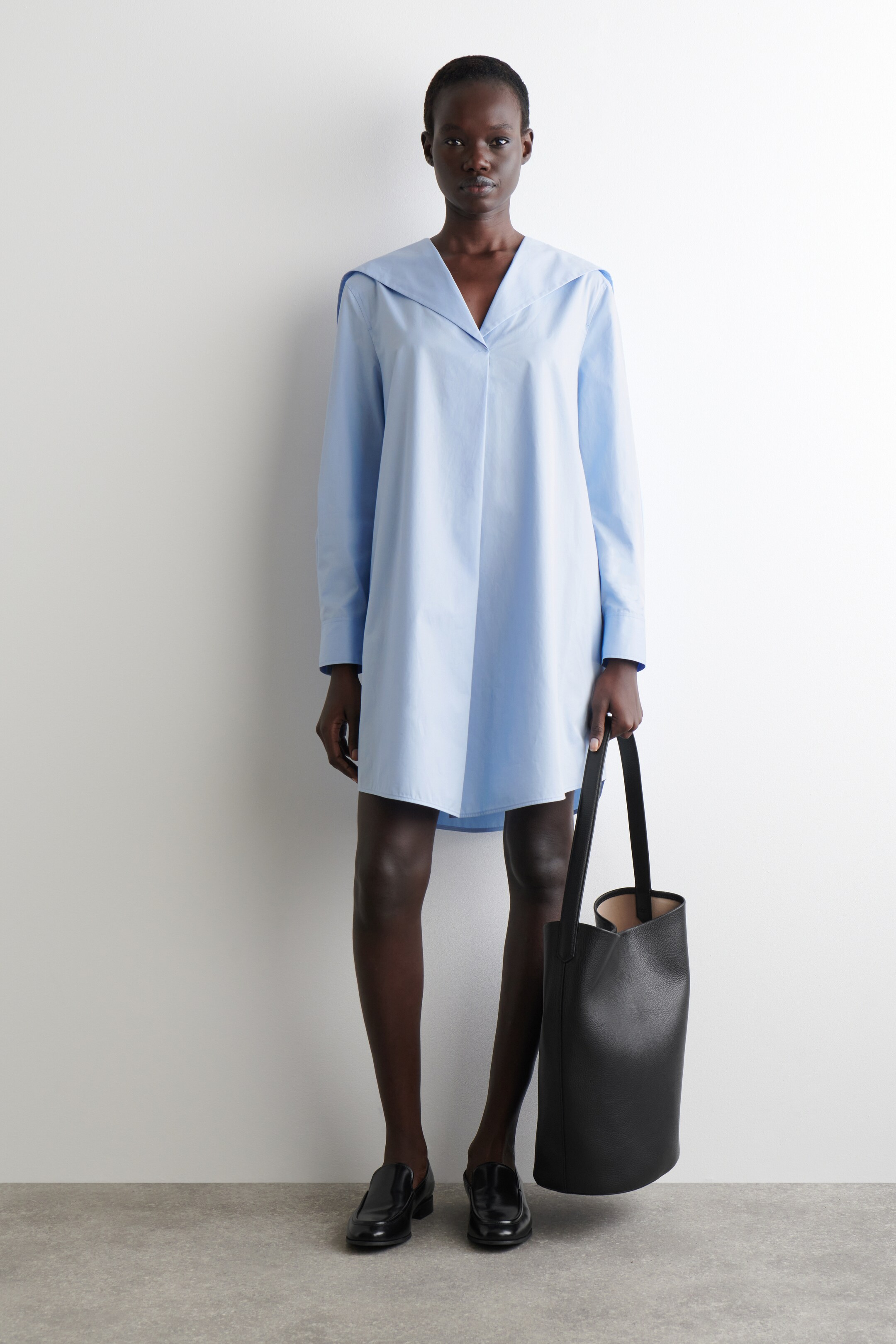 Agrandir l'image: Une robe-chemise bleu clair est présentée, avec un large col pointu qui s'étend sur les épaules et forme un profond décolleté en V. La robe présente une coupe ample et décontractée avec des manches longues et un ourlet au-dessus du genou.
