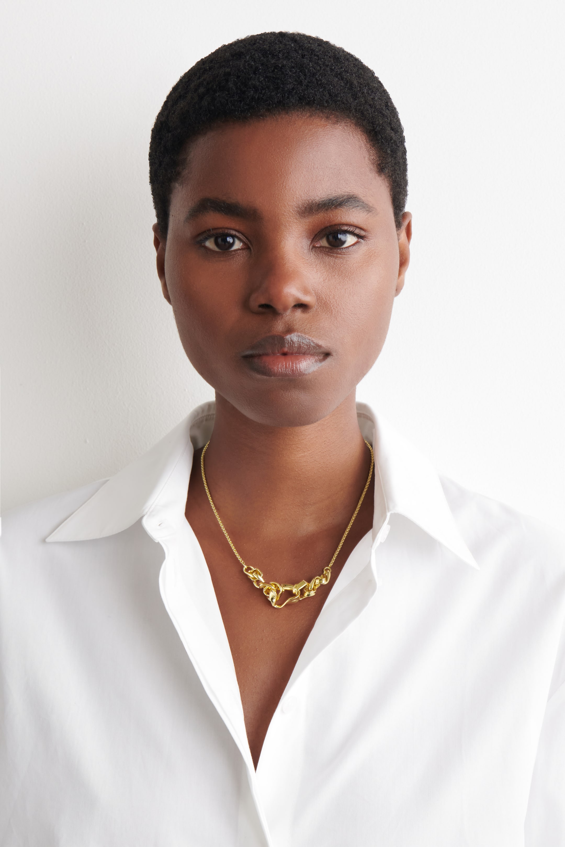 הצגת תמונה גדולה יותר: שרשרת בגוון זהב עם חלק מרכזי בולט של חוליות לא סדירות המשתלבות זו בזו, עם שרשראות דקות יותר המשתרעות מכל צד. היא מונחת מתחת לעצמות הבריח מעל חולצה לבנה עם צווארון פתוח, פונה קדימה.