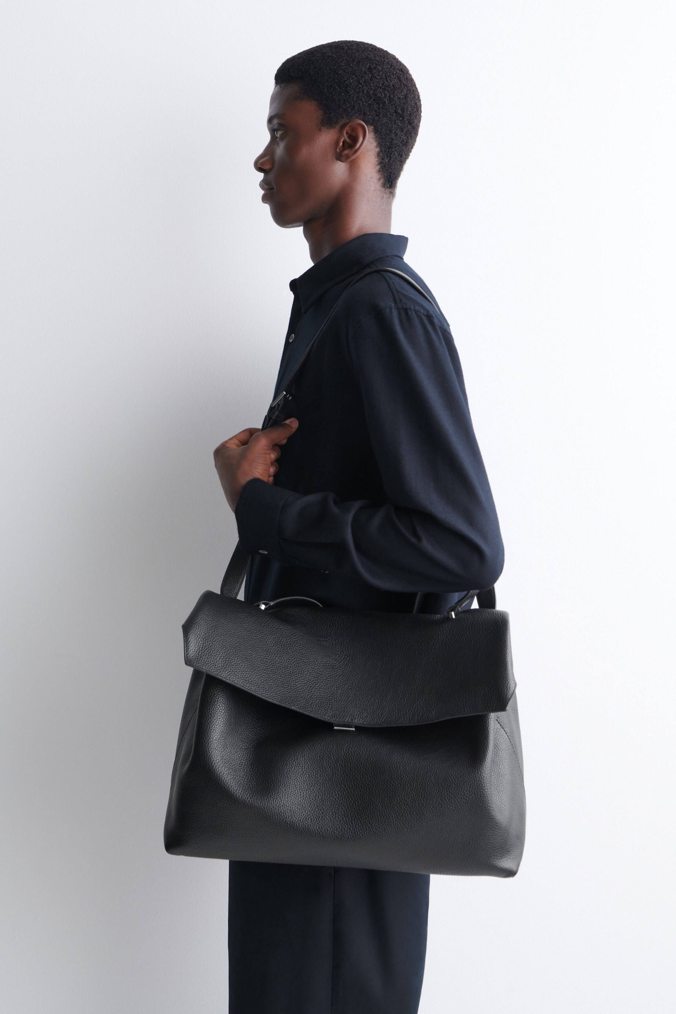El bolso Trullo oversize con solapa, en cuero negro granuloso, lo lleva la modelo colgado del hombro mientras sujeta la correa. El bolso presenta una gran solapa frontal, un pequeño asa superior y un acabado texturizado. El bolso Trullo oversize con solapa, en cuero negro granuloso, lo lleva la modelo colgado del hombro mientras sujeta la correa. El bolso presenta una gran solapa frontal, un pequeño asa superior y un acabado texturizado.