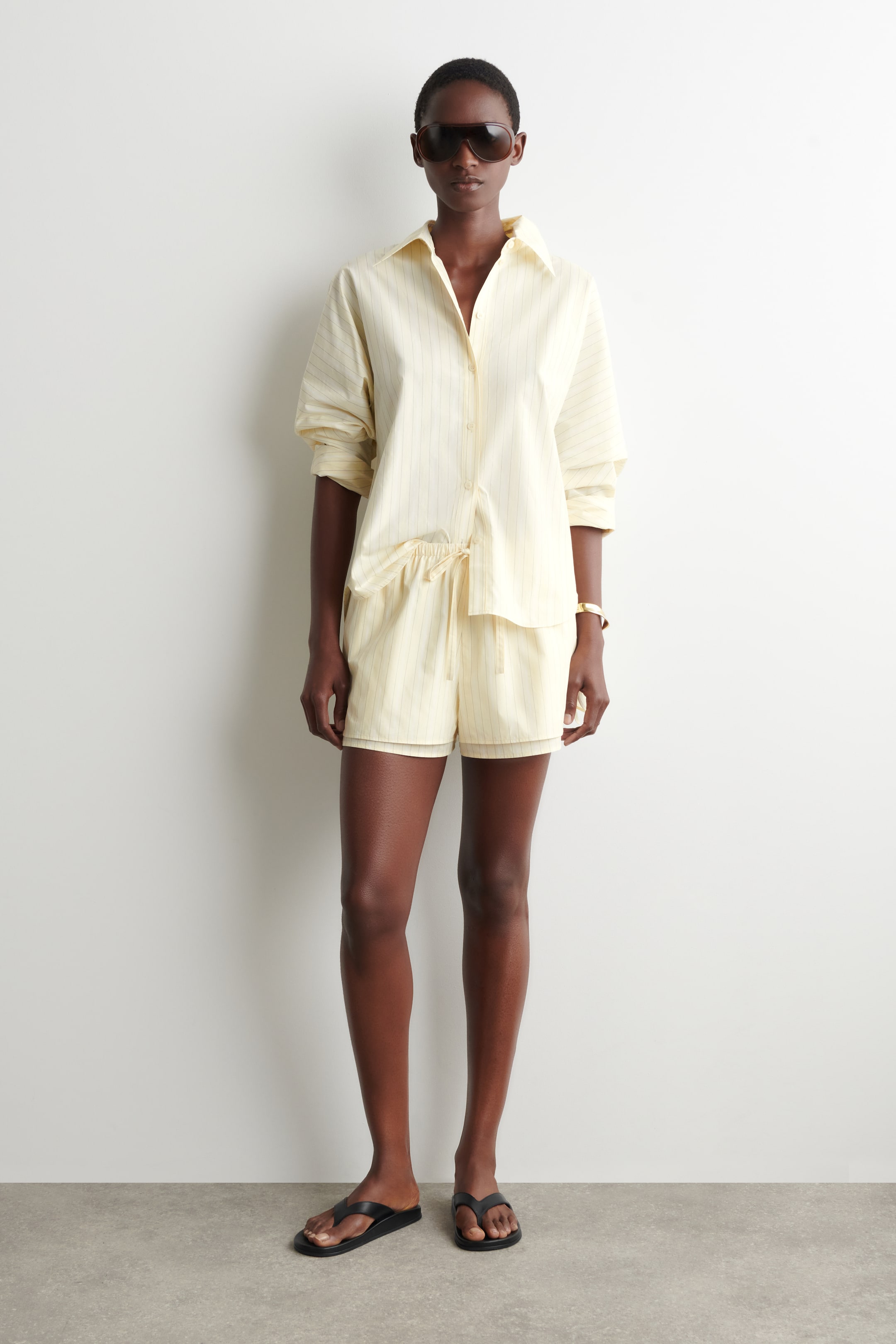 Visualizza immagine più grande: Una camicia a righe sottili giallo chiaro con bottoni e pantaloncini con coulisse abbinati sono in mostra. La camicia presenta un colletto e maniche lunghe arrotolate, e la parte anteriore è parzialmente infilata nei pantaloncini.