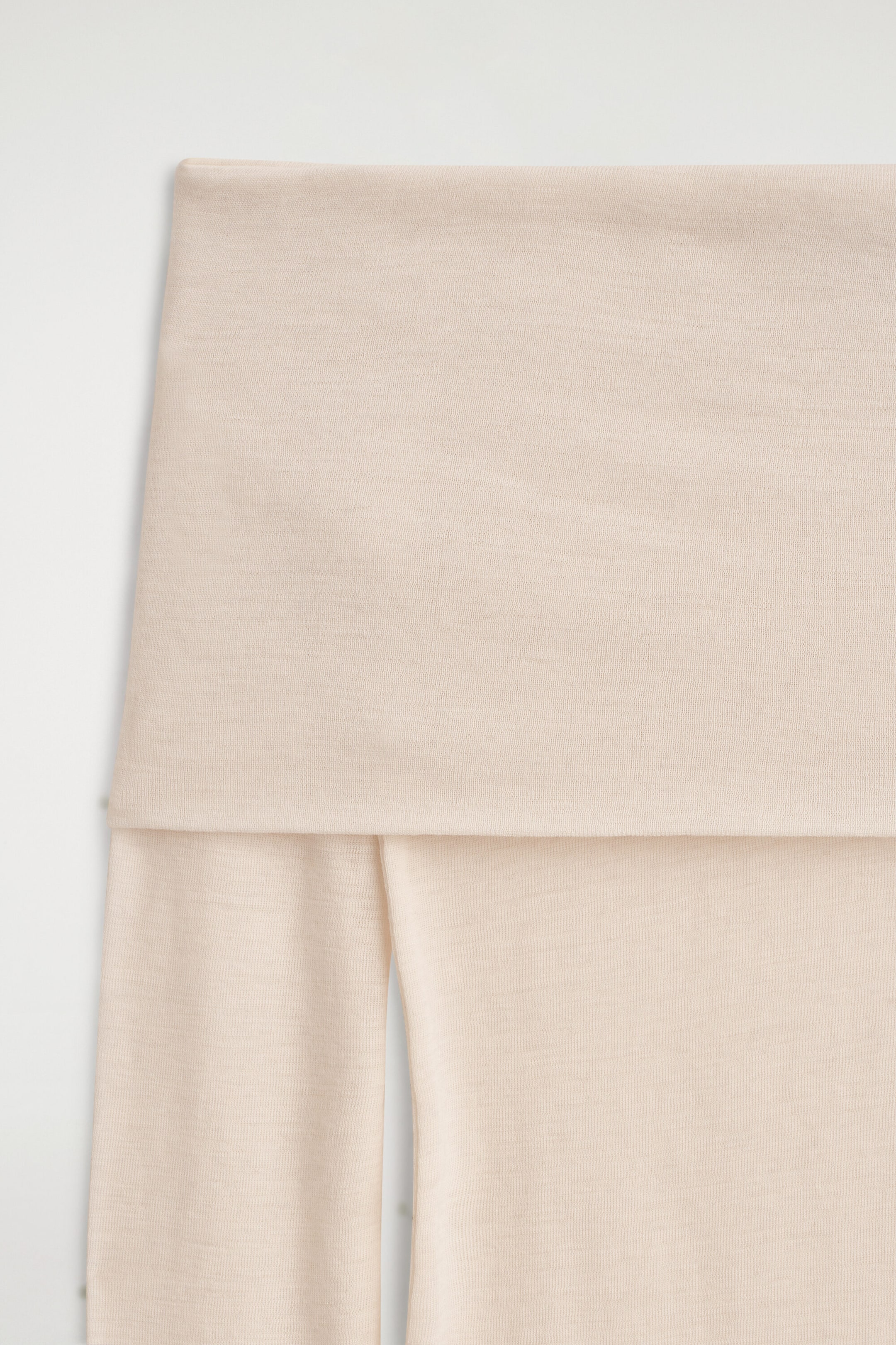 A light beige off-the-shoulder merino wool top is shown in a close-up front view, highlighting its fine-knit texture. The garment features a wide, straight foldover neckline that drapes over the bodice and long sleeves. A light beige off-the-shoulder merino wool top is shown in a close-up front view, highlighting its fine-knit texture. The garment features a wide, straight foldover neckline that drapes over the bodice and long sleeves.