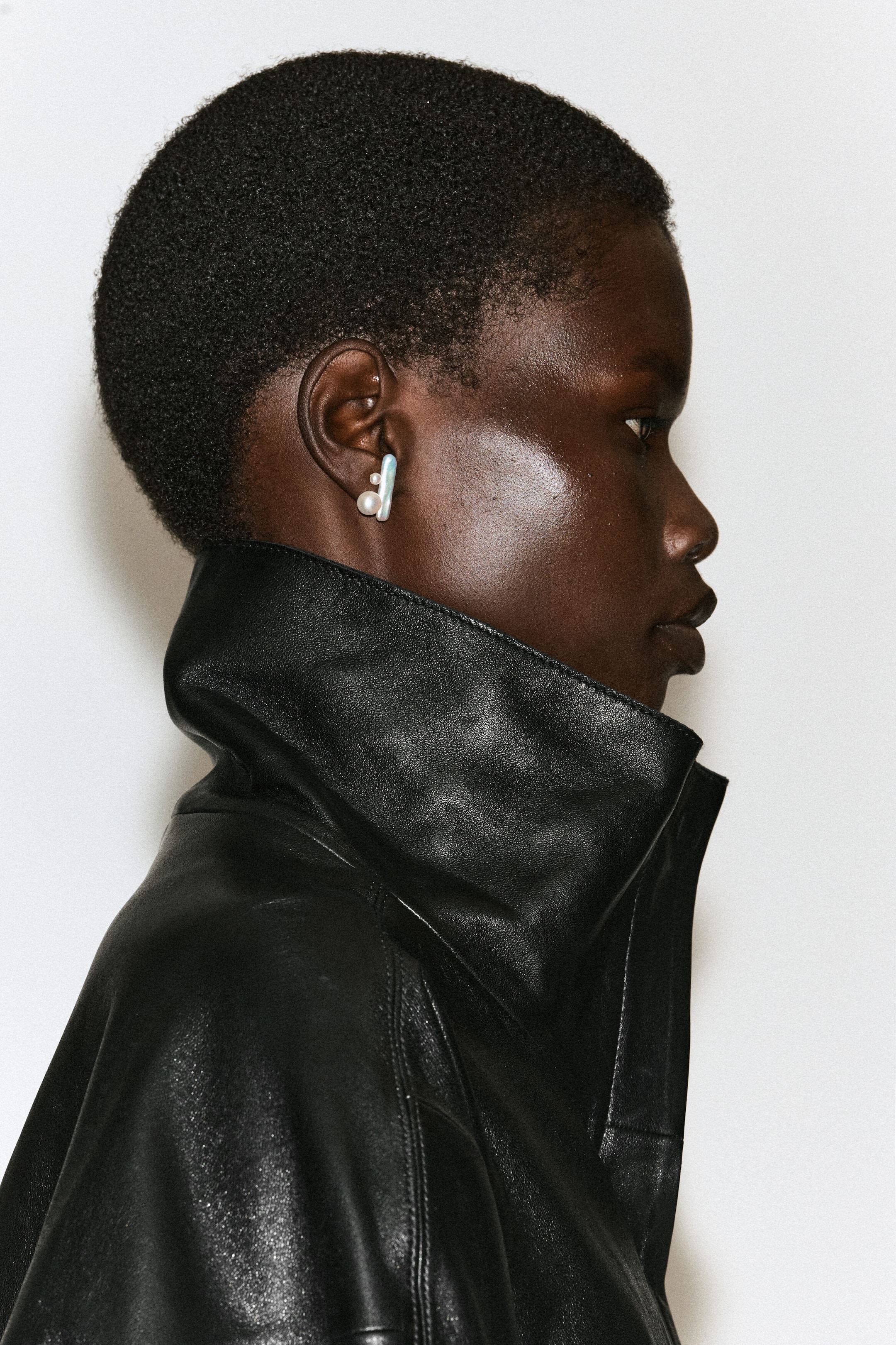 Sculptural freshwater pearl earrings, featuring a vertical bar-shaped pearl and a smaller round pearl, are being worn by the model. The earring is styled alongside a high-collared black leather jacket. Sculptural freshwater pearl earrings, featuring a vertical bar-shaped pearl and a smaller round pearl, are being worn by the model. The earring is styled alongside a high-collared black leather jacket.