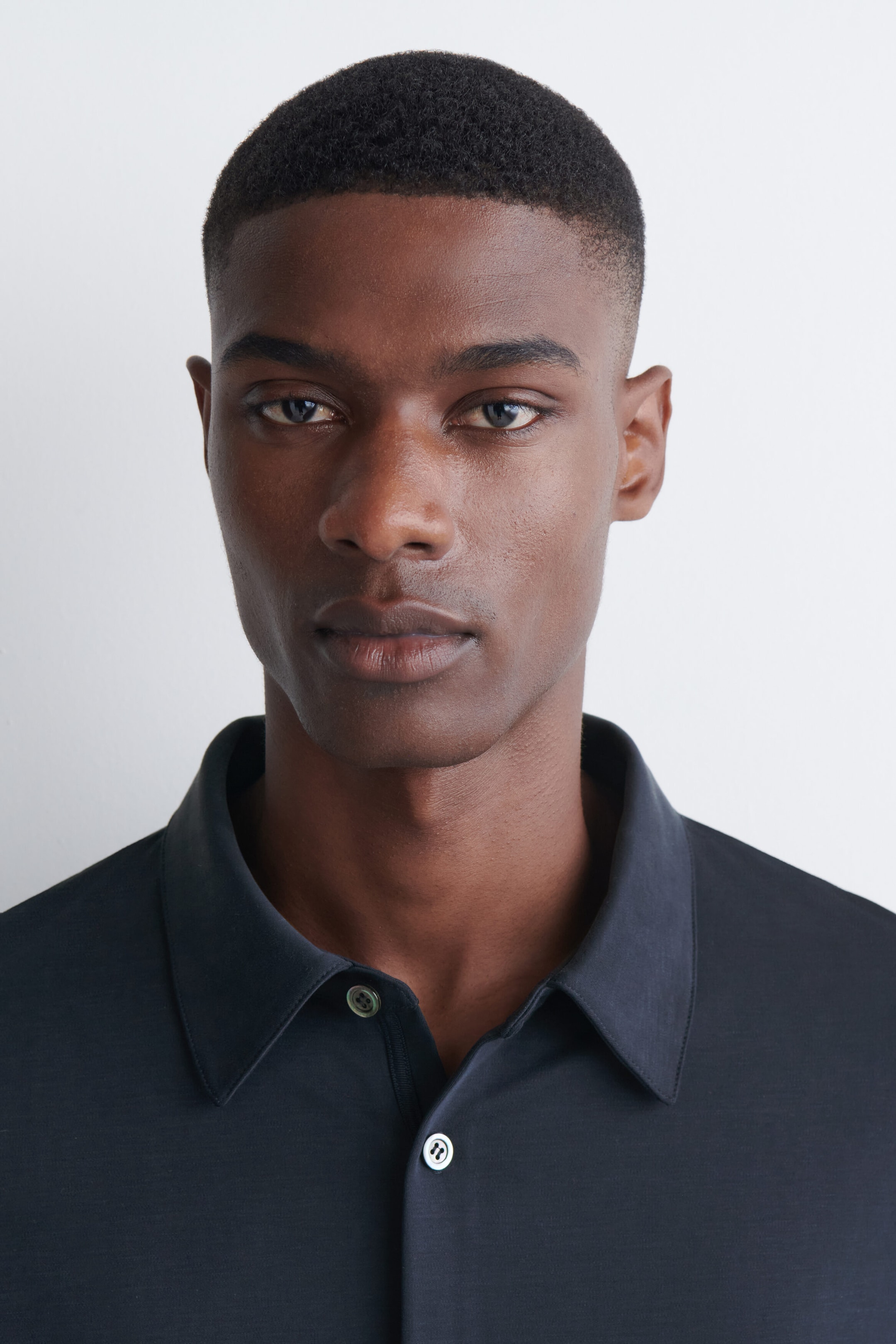 The model is wearing a dark gray fluid interlock jersey shirt featuring a classic point collar and a partially unbuttoned placket with light-colored buttons. The model is wearing a dark gray fluid interlock jersey shirt featuring a classic point collar and a partially unbuttoned placket with light-colored buttons.