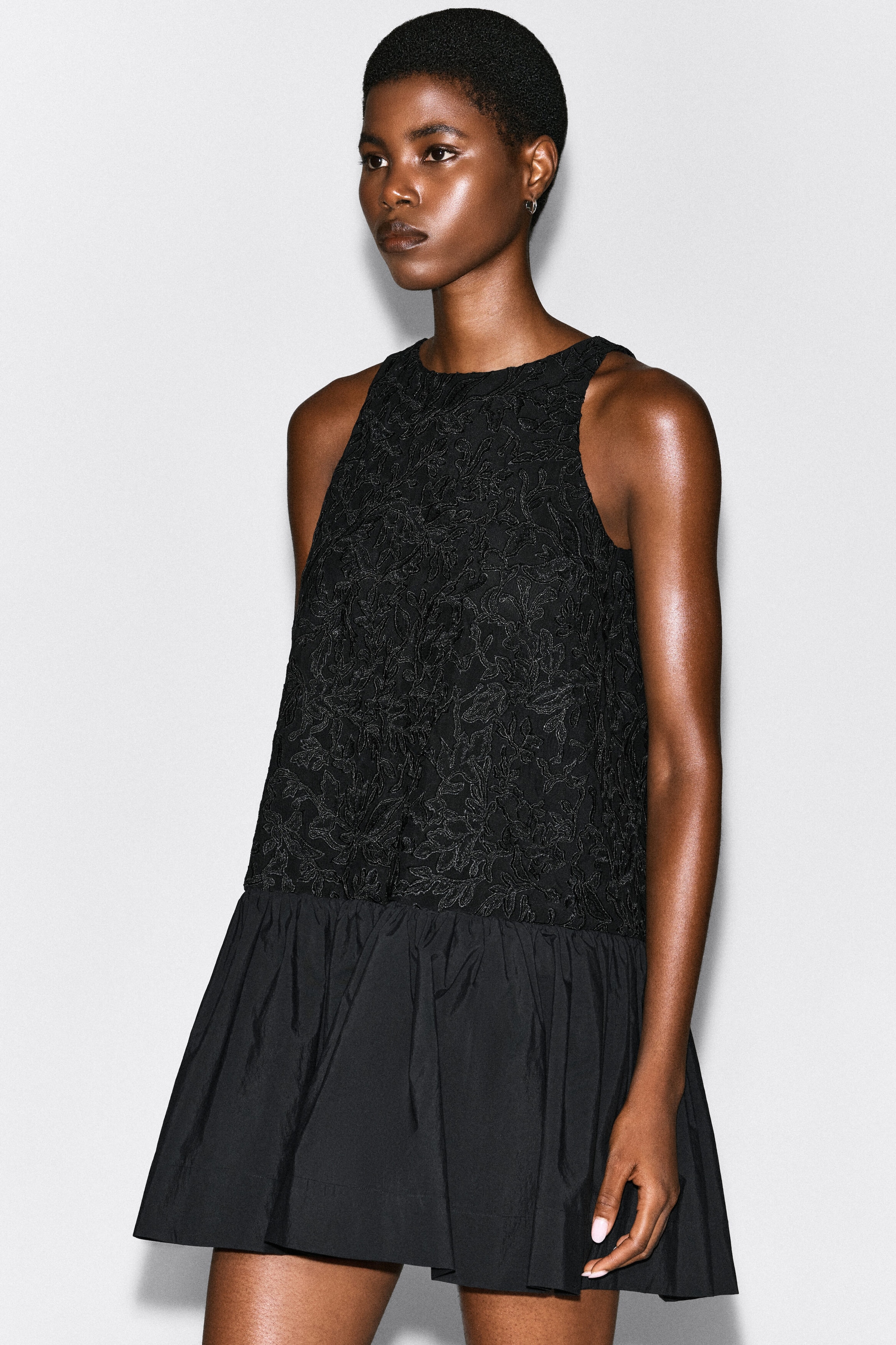 The model is wearing a sleeveless black dress featuring an intricately embroidered floral lace bodice and a contrasting, voluminous poplin balloon skirt. The model is wearing a sleeveless black dress featuring an intricately embroidered floral lace bodice and a contrasting, voluminous poplin balloon skirt.