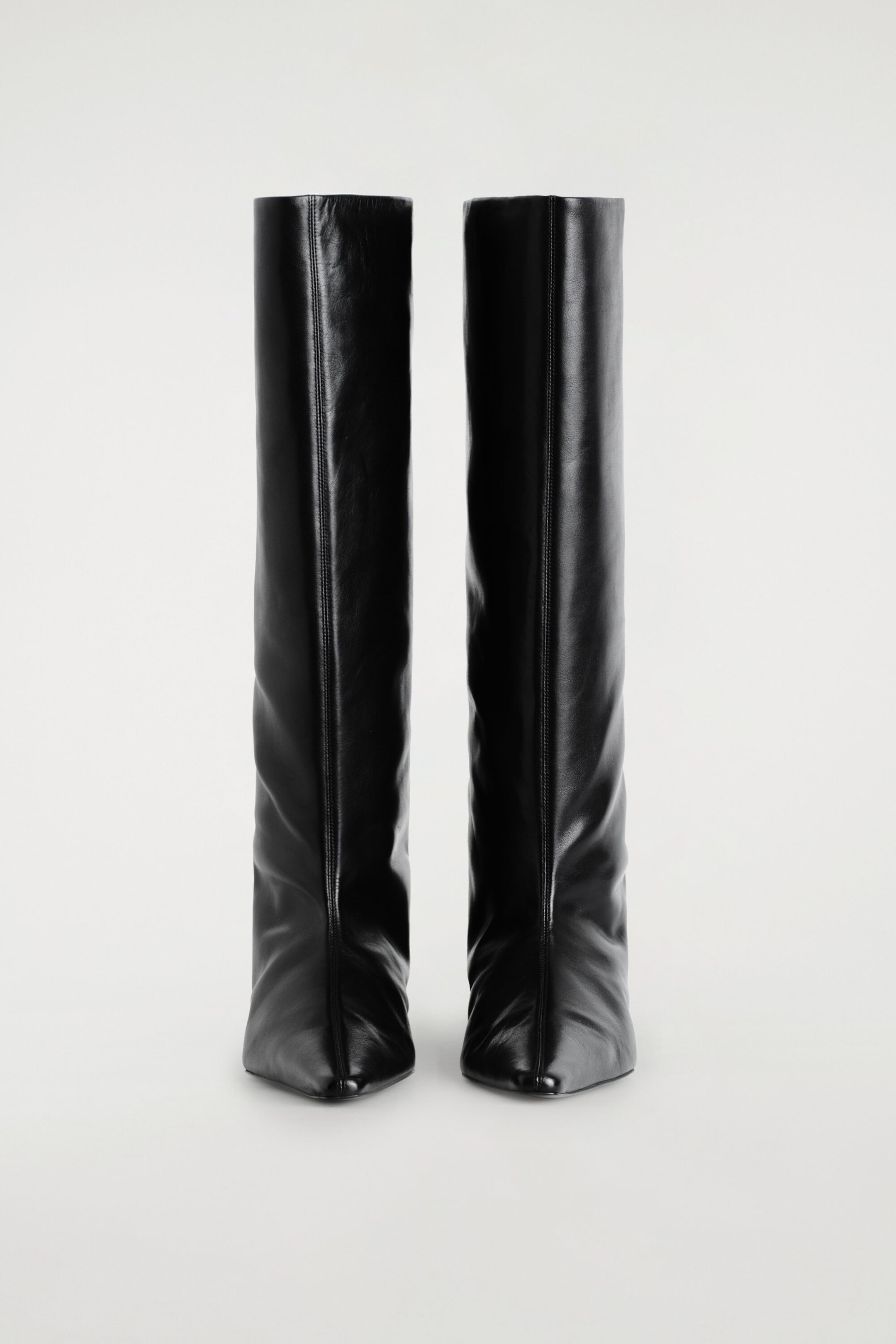A pair of black leather knee-high boots is positioned front-facing, featuring a sharp pointed toe and a prominent vertical seam running down the center of each straight, wide shaft. The boots showcase a smooth, structured leather design with subtle creasing near the ankles. A pair of black leather knee-high boots is positioned front-facing, featuring a sharp pointed toe and a prominent vertical seam running down the center of each straight, wide shaft. The boots showcase a smooth, structured leather design with subtle creasing near the ankles.