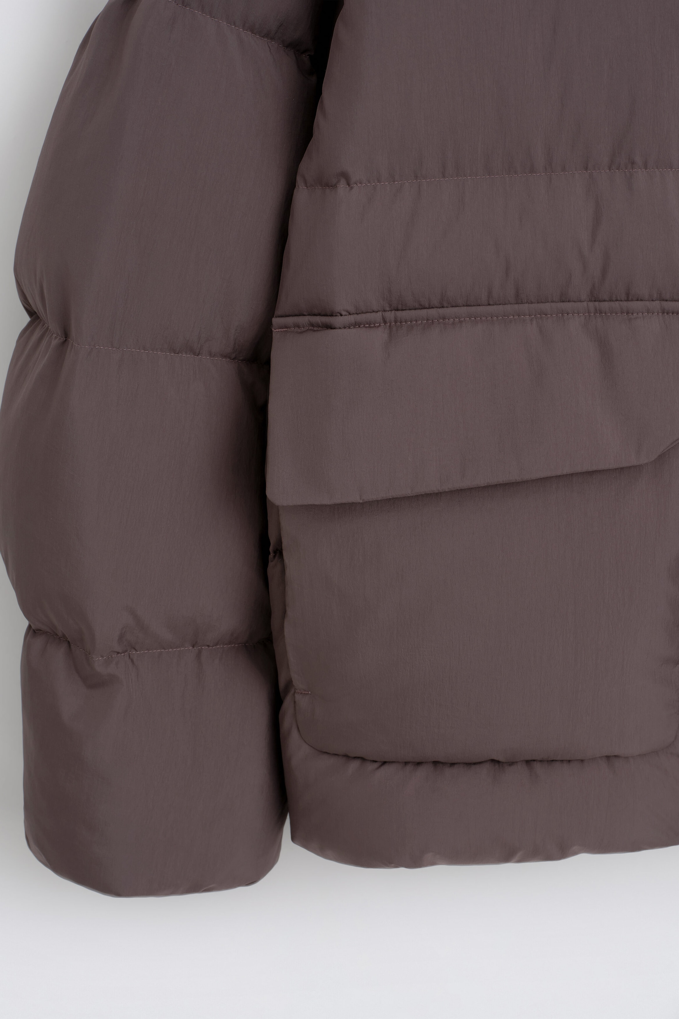 A close-up of the funnel-neck nylon down puffer jacket in dark brown shows its horizontal quilting and a large front flap pocket. The matte fabric is constructed with thick, insulated puffy panels on both the body and sleeves. A close-up of the funnel-neck nylon down puffer jacket in dark brown shows its horizontal quilting and a large front flap pocket. The matte fabric is constructed with thick, insulated puffy panels on both the body and sleeves.