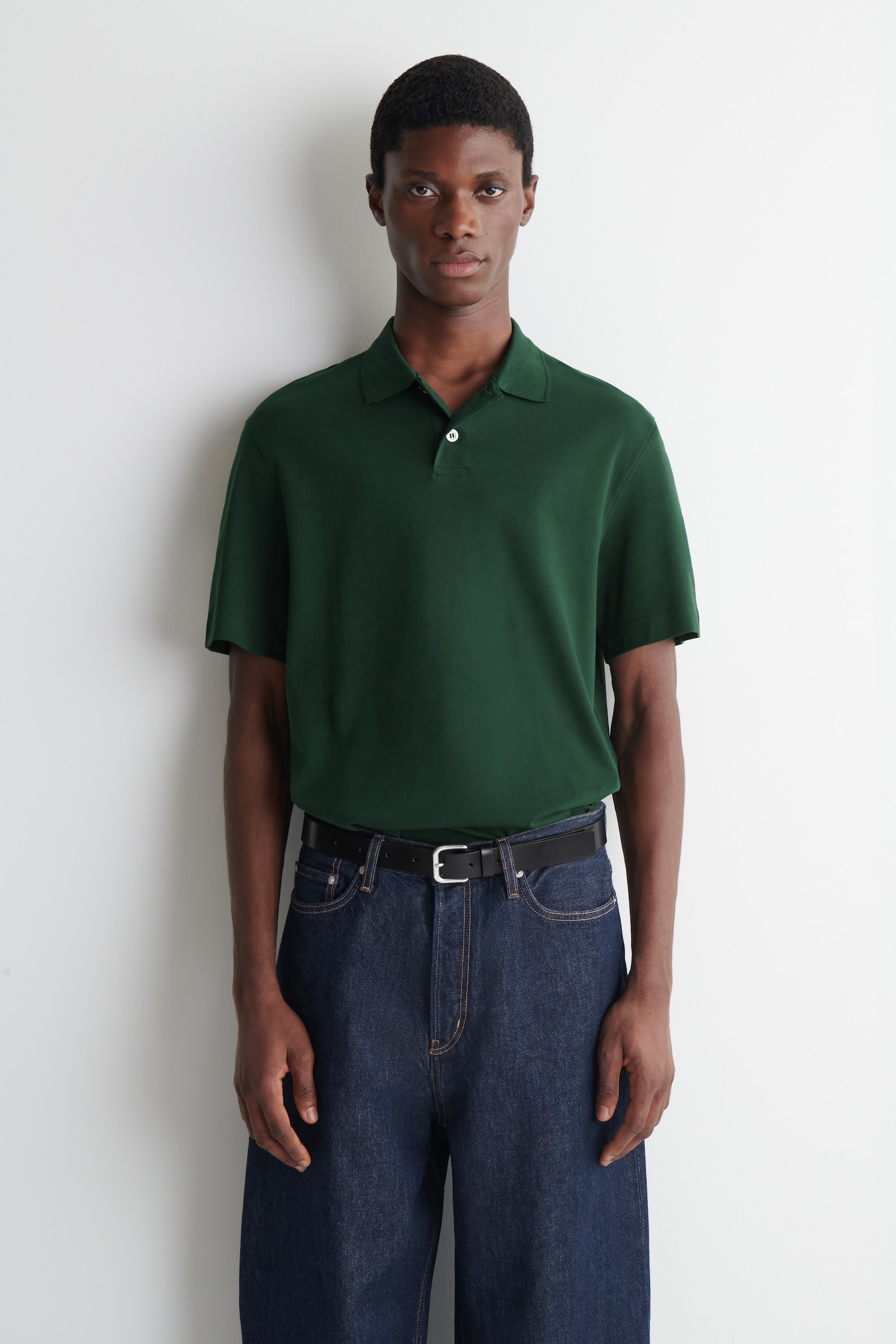 Een donkergroen poloshirt met korte mouwen en een klassieke kraag met een gedeeltelijk geopende knoopsluiting wordt gedragen door het model. Het shirt is ingestopt in donkerblauwe wijde broekspijpen. - 1