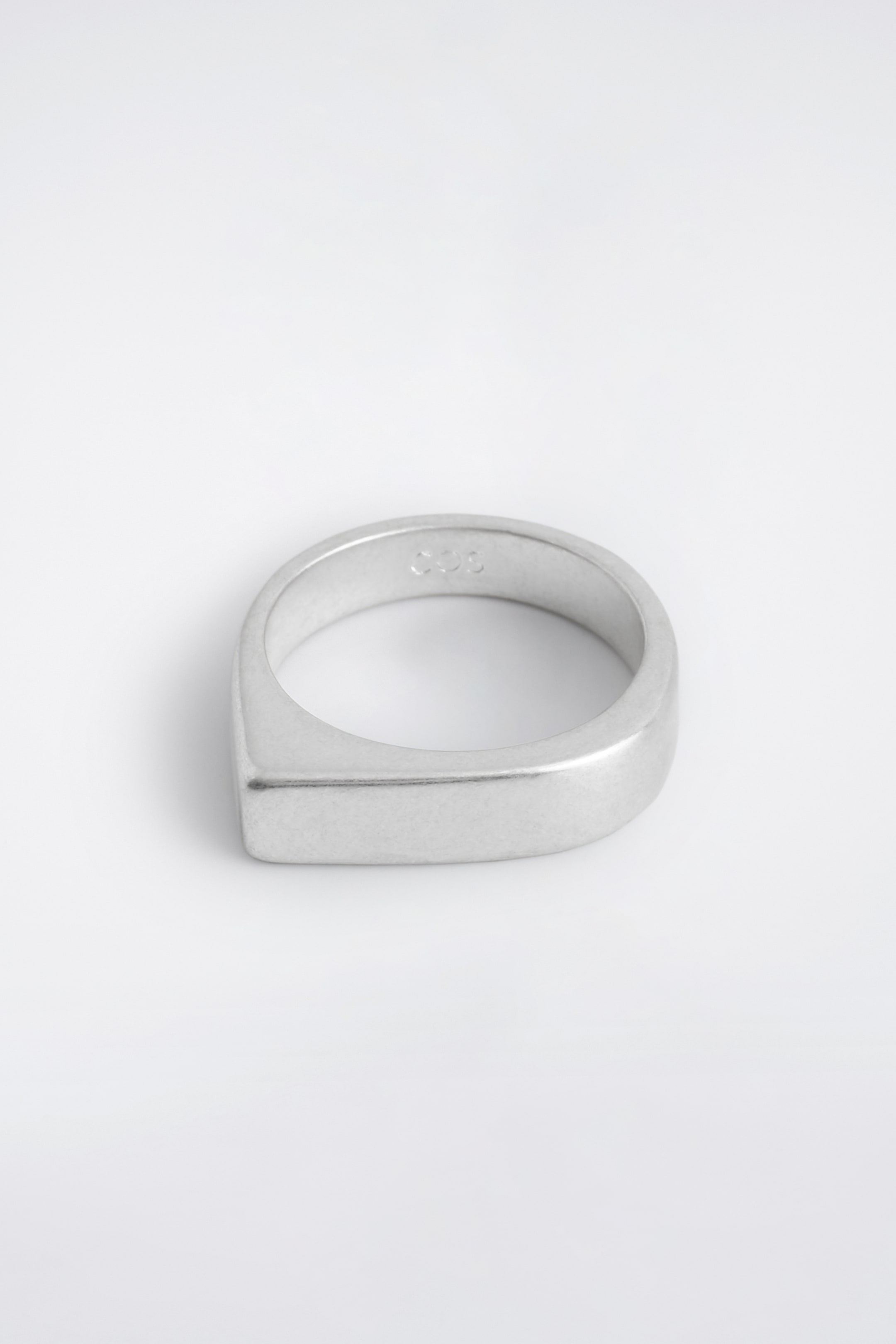 Visualizza immagine più grande: Un anello con sigillo placcato in argento presenta un design scultoreo, con la sua superficie superiore che si curva da un lato e si squadrata dall'altro. L'anello presenta una finitura opaca, leggermente ossidata.