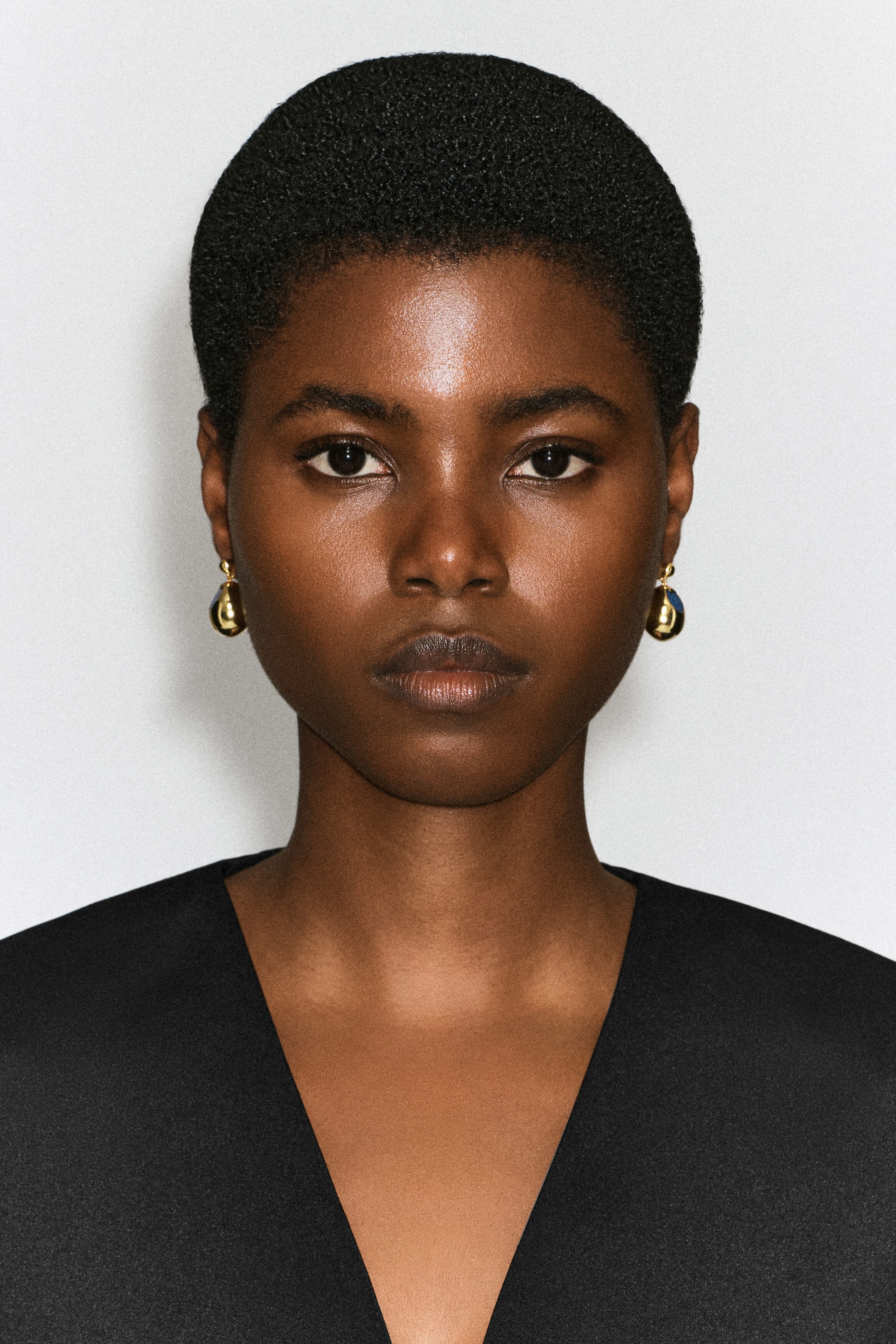Des boucles d’oreilles pendantes ton or, de forme arrondie et bombée, ornées d’un petit détail en pierre bleue, sont portées par le modèle. Les boucles d’oreilles polies sont portées avec un haut noir à col en V.