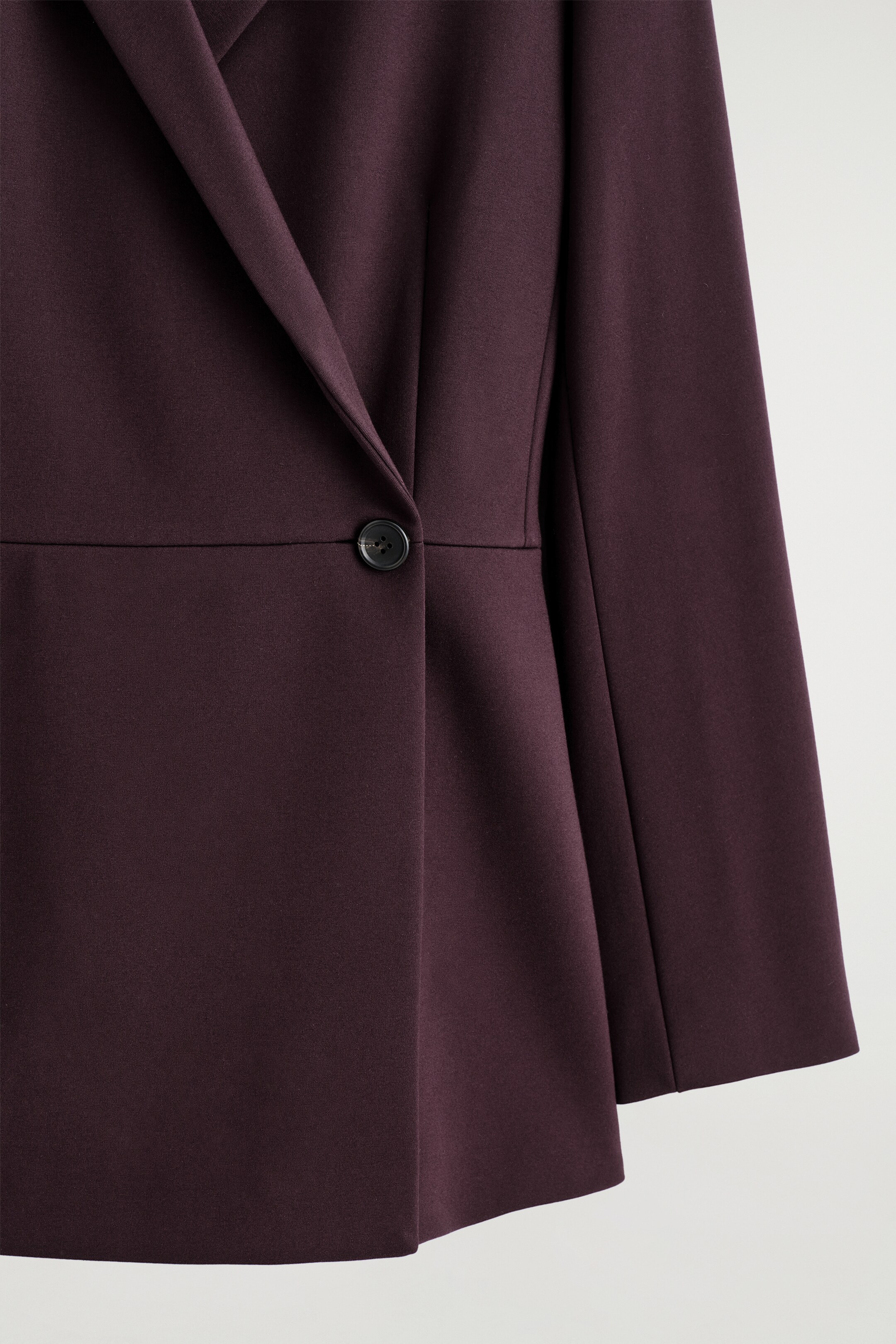 Visualizza immagine più grande: Un primo piano di un blazer bordeaux scuro, caratterizzato da una silhouette a portafoglio. Un singolo bottone scuro chiude il davanti e una cucitura orizzontale definisce la linea della vita.