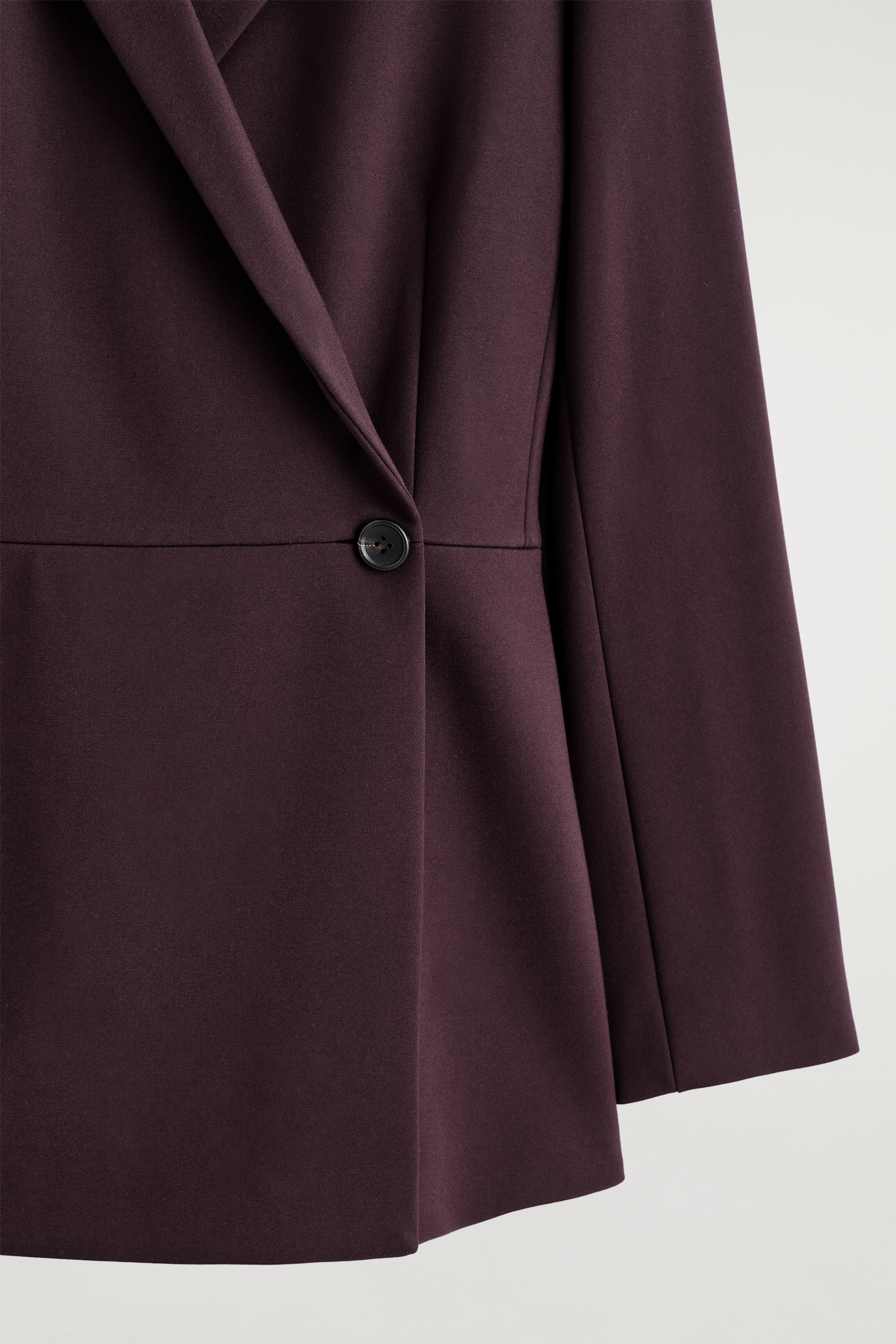Visualizza immagine più grande: Un primo piano di un blazer bordeaux scuro, caratterizzato da una silhouette a portafoglio. Un singolo bottone scuro chiude il davanti e una cucitura orizzontale definisce la linea della vita.