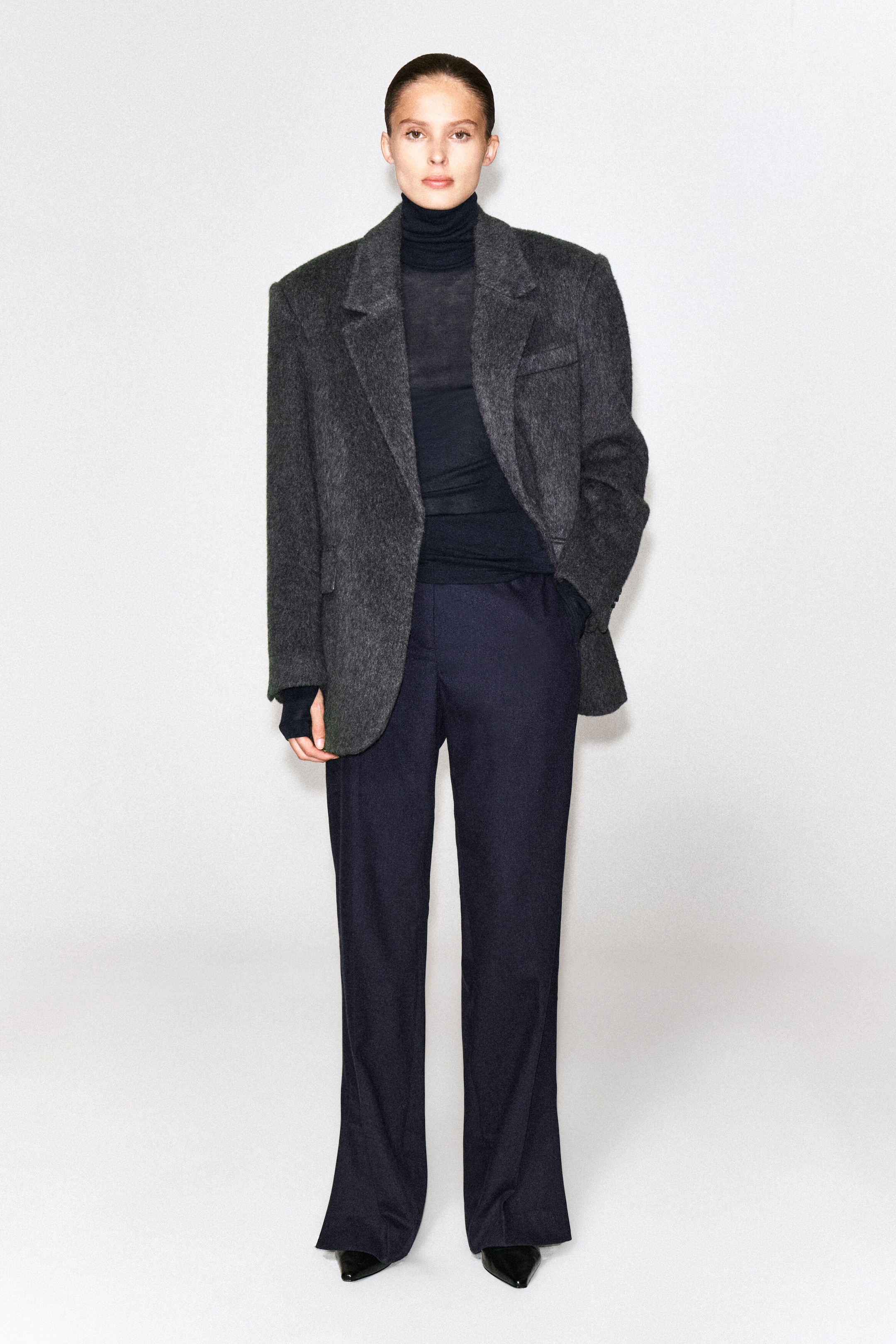 A merino wool high-neck top in a dark shade is worn by the model as a base layer under a textured gray blazer and tucked into dark navy trousers. The fine-knit top features a high, fitted collar and long sleeves that extend past the blazer cuffs.